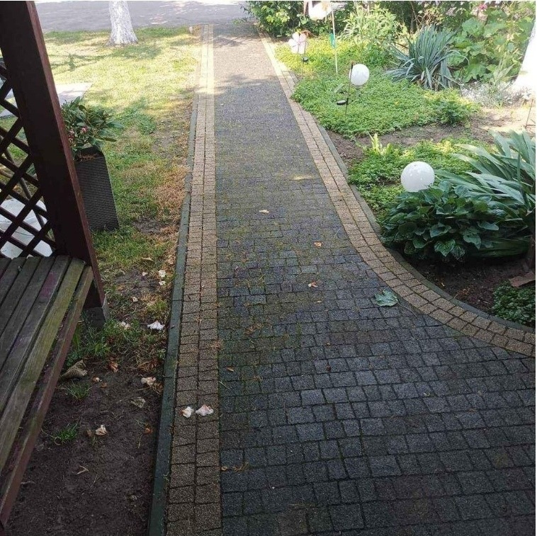 Ciemnoszara kostka brukowa z brązową obwódką, częściowo pokryta mchem i liśćmi, widok z góry, fragment drewnianej konstrukcji po lewej stronie.