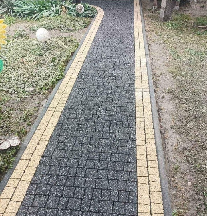 Ścieżka z ciemnoszarej kostki brukowej z jasnymi obrzeżami, prowadząca przez ogród z ozdobnymi kulami i roślinnością.