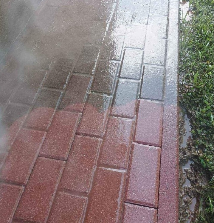Częściowo umyta, mokra kostka brukowa w odcieniach czerwieni, z widoczną różnicą czystości między umytą i nieumytą powierzchnią, oraz fragment trawnika po prawej stronie.
