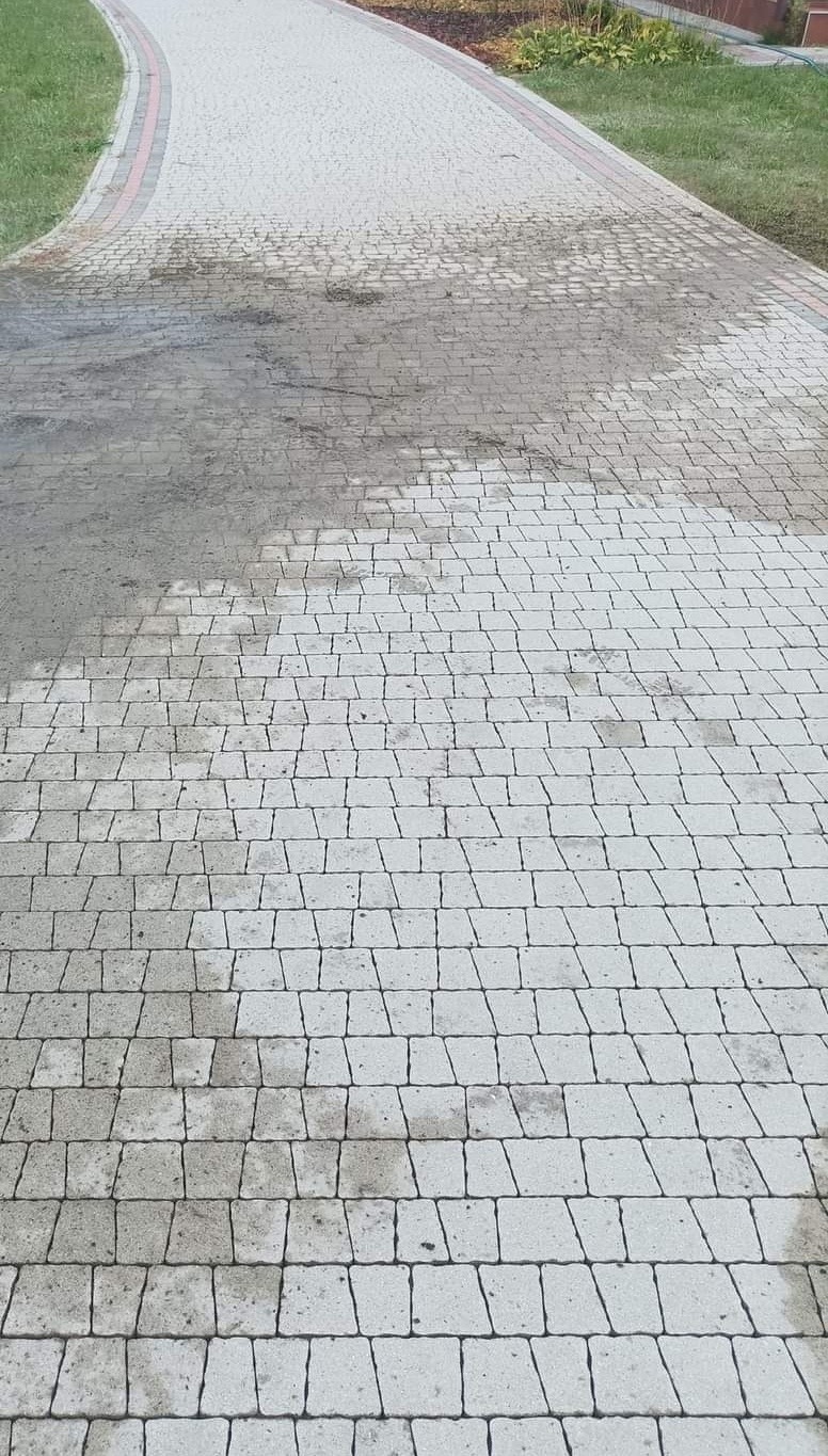 Częściowo wyczyszczona kostka brukowa na zakrzywionej ścieżce, widoczne wyraźne różnice w kolorze między brudną a czystą powierzchnią, trawnik po obu stronach.
