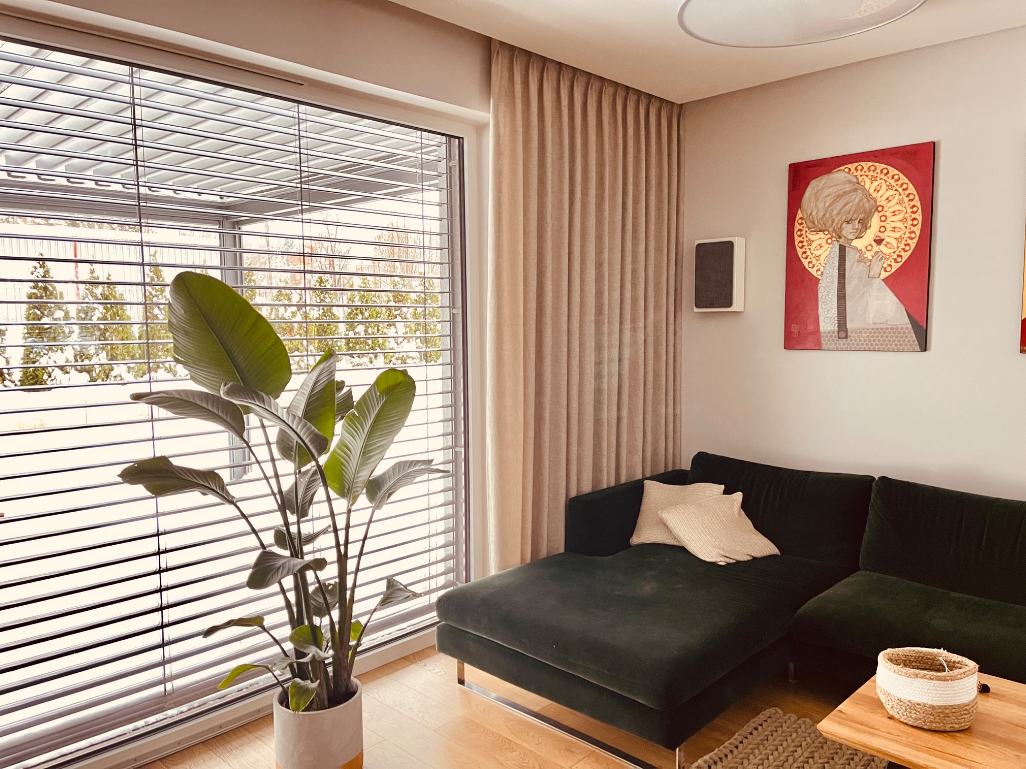 Nowoczesny salon z zieloną sofą, rośliną doniczkową przy oknie z poziomymi żaluzjami i beżowymi zasłonami, uzupełniony obrazem na ścianie.