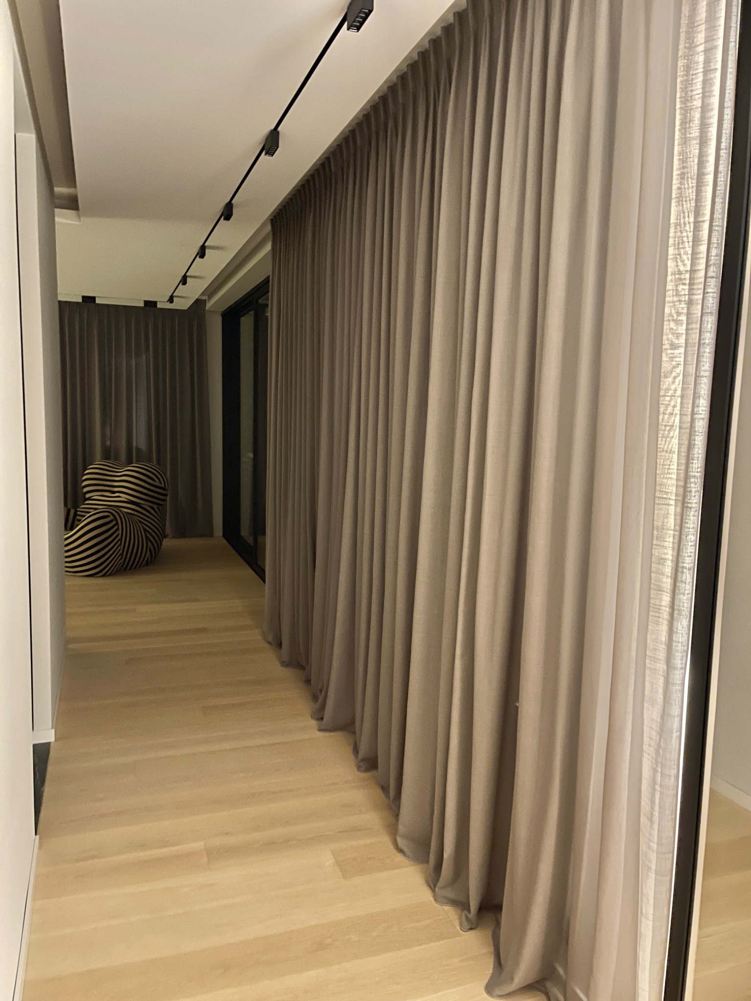 Długie, beżowe zasłony w nowoczesnym wnętrzu, z widocznym systemem szynowym pod sufitem i designerskim fotelem w tle.