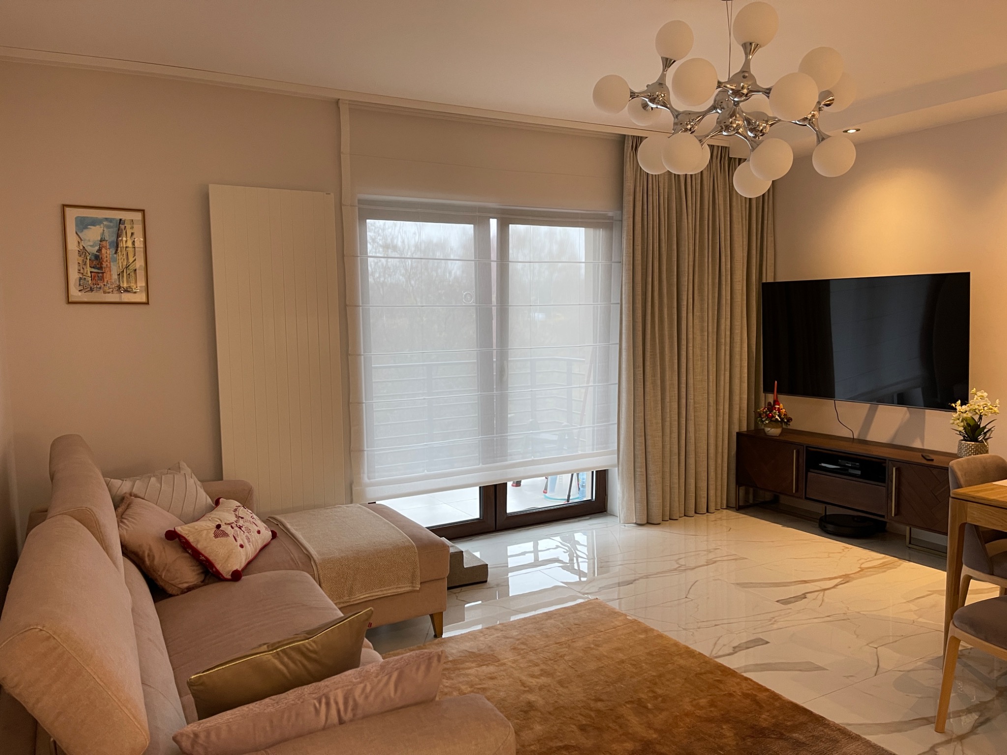 Jasne, nowoczesne wnętrze salonu z marmurową podłogą, beżową sofą z poduszkami, telewizorem na komodzie, oraz roletami rzymskimi w oknie i zasłonami po bokach okna.