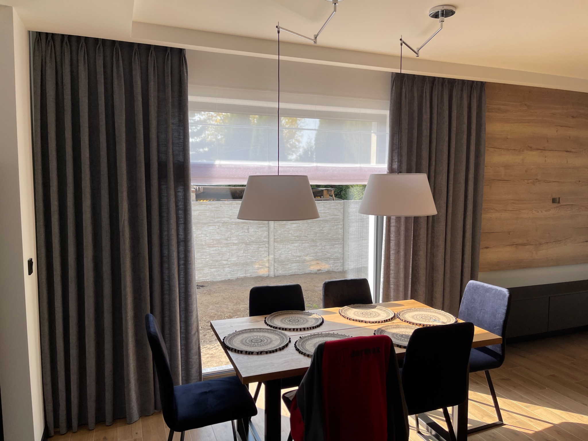 Nowoczesna jadalnia z drewnianym stołem, czarnymi krzesłami i szarymi zasłonami z delikatnym marszczeniem, oświetlona dwoma lampami wiszącymi z białymi abażurami. W oknie widoczna biała roleta...