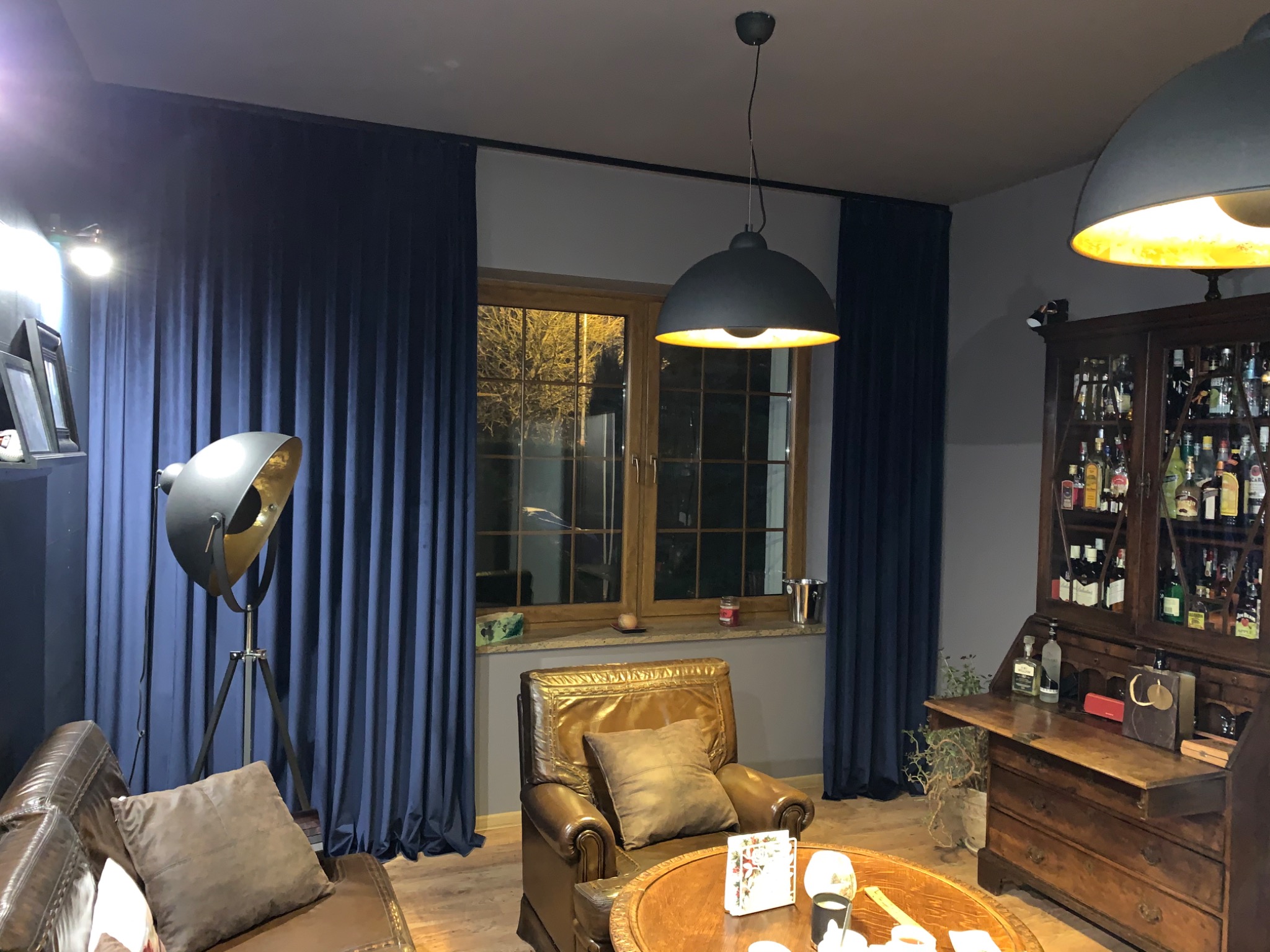 Eleganckie wnętrze z granatowymi zasłonami, lampą studyjną na trójnogu, skórzanym fotelem i barkiem z alkoholem, oświetlone ciepłym światłem wiszących lamp.