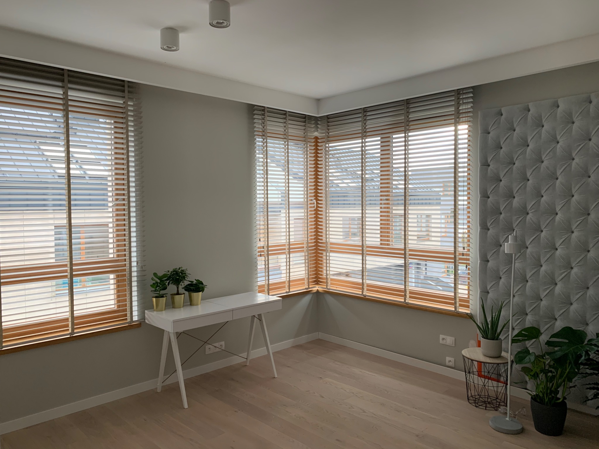 Jasne wnętrze z drewnianymi żaluzjami poziomymi w oknach, biurko z roślinami w złotych doniczkach, pikowany panel ścienny i drewniana podłoga.