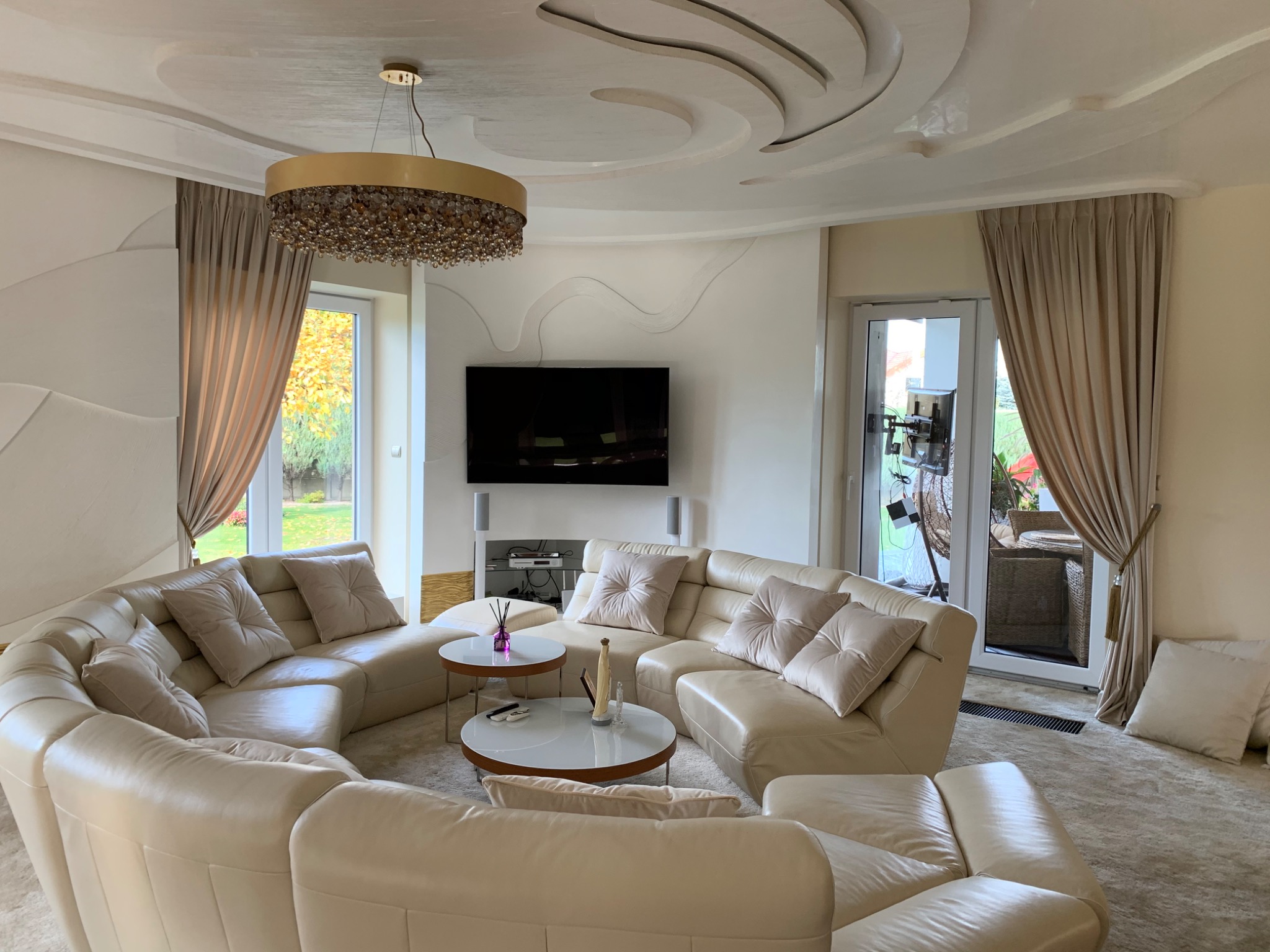 Elegancki salon z beżową, modułową sofą w kształcie podkowy, okrągłymi stolikami kawowymi, beżowymi zasłonami z dekoracyjnymi chwostami i nowoczesnym oświetleniem wiszącym.