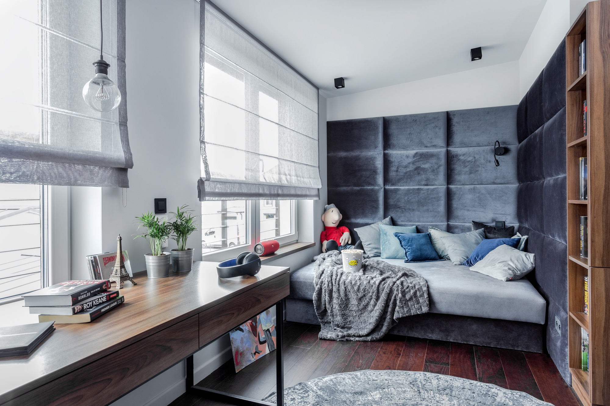 Nowoczesny pokój z szarymi roletami rzymskimi, drewnianym biurkiem z dekoracjami i tapicerowanym panelem ściennym w kolorze grafitowym.