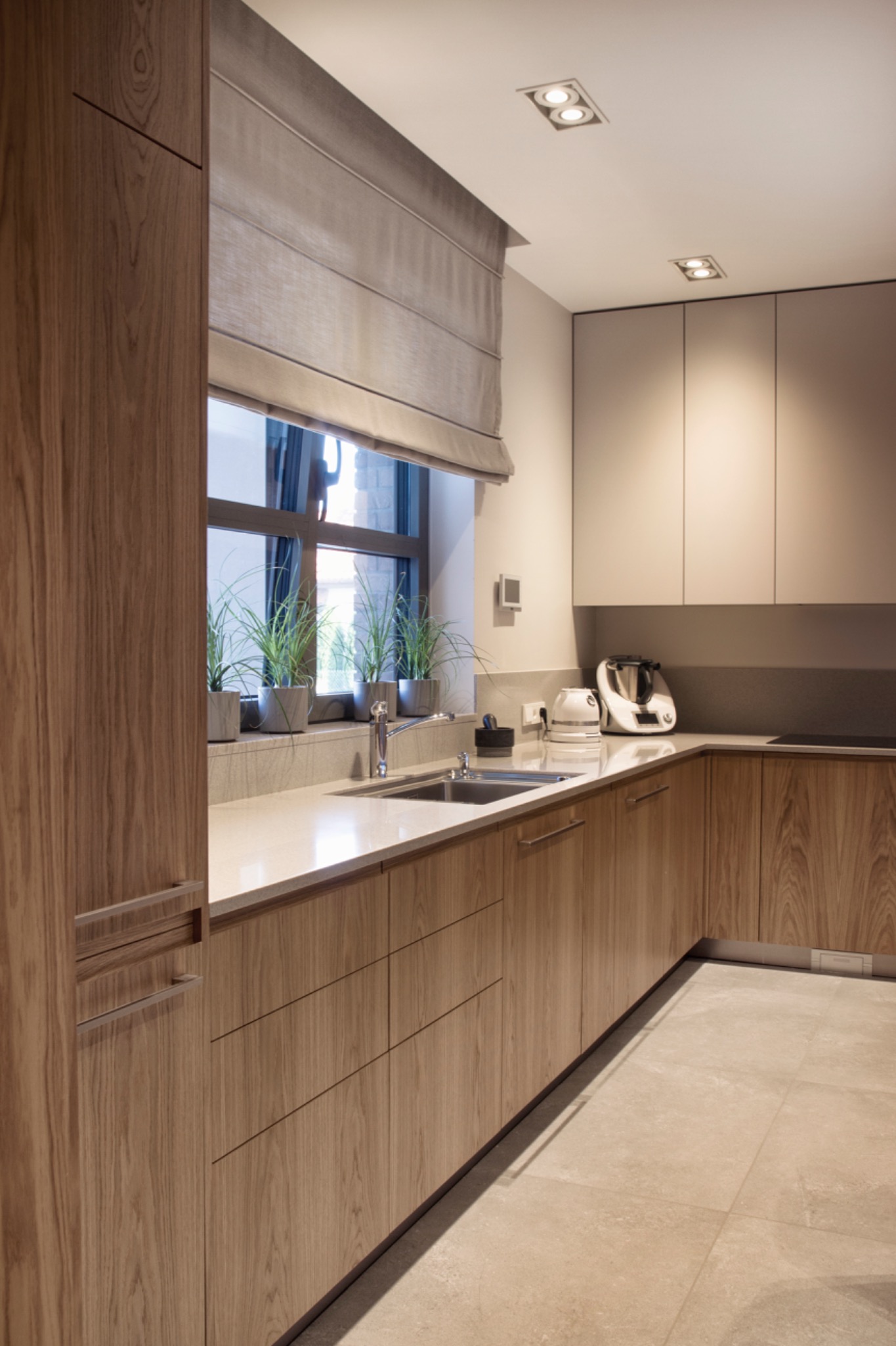 Jasna kuchnia z drewnianymi szafkami, blatem z roślinami w doniczkach przy oknie z szarą, zwiniętą roletą rzymską i nowoczesnym sprzętem AGD.