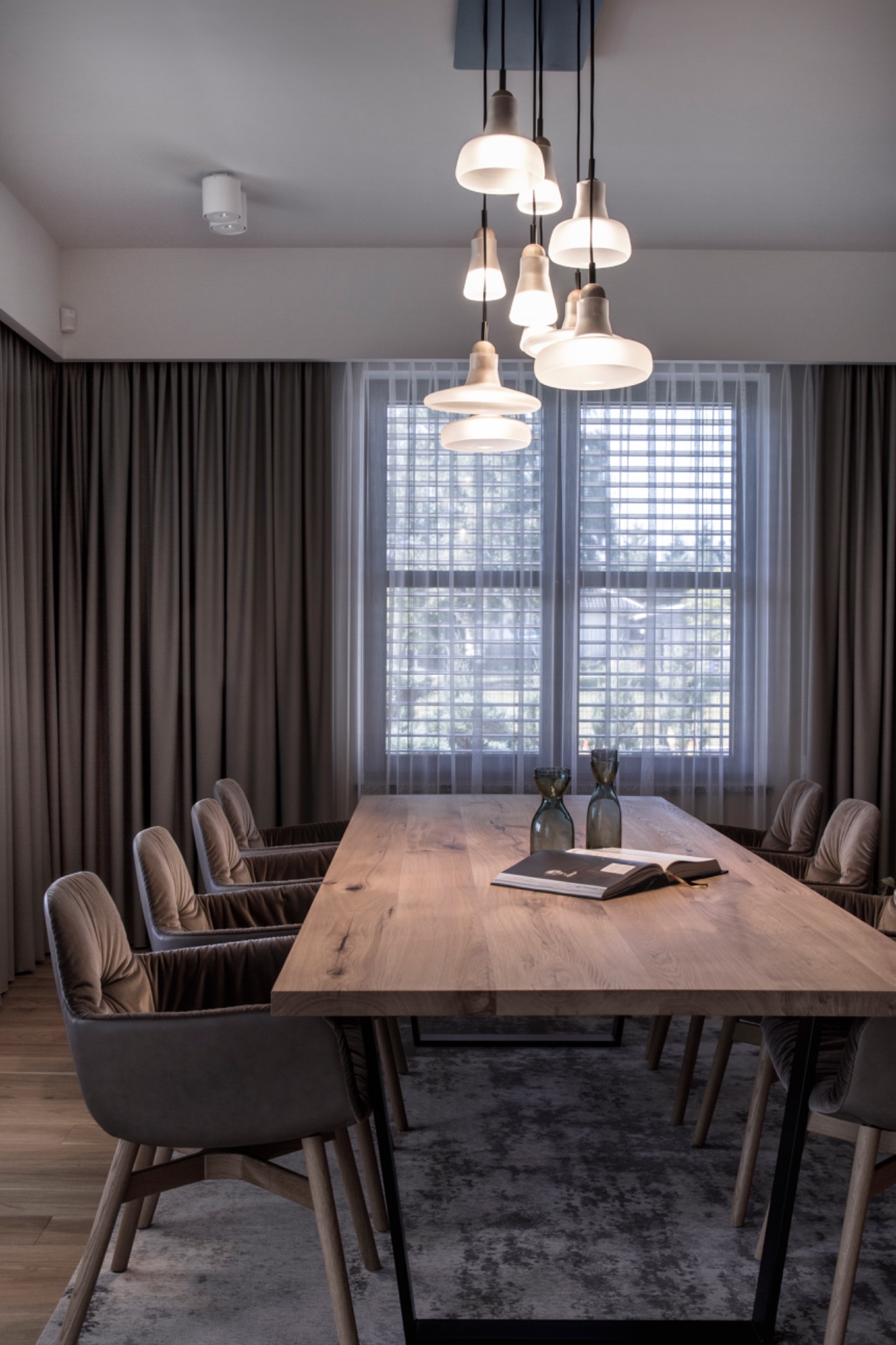 Jadalnia z długim drewnianym stołem, krzesłami z tapicerowanymi oparciami, szarymi zasłonami po bokach okna z delikatną, białą roletą w kratkę i wiszącymi lampami o różnej wysokości.