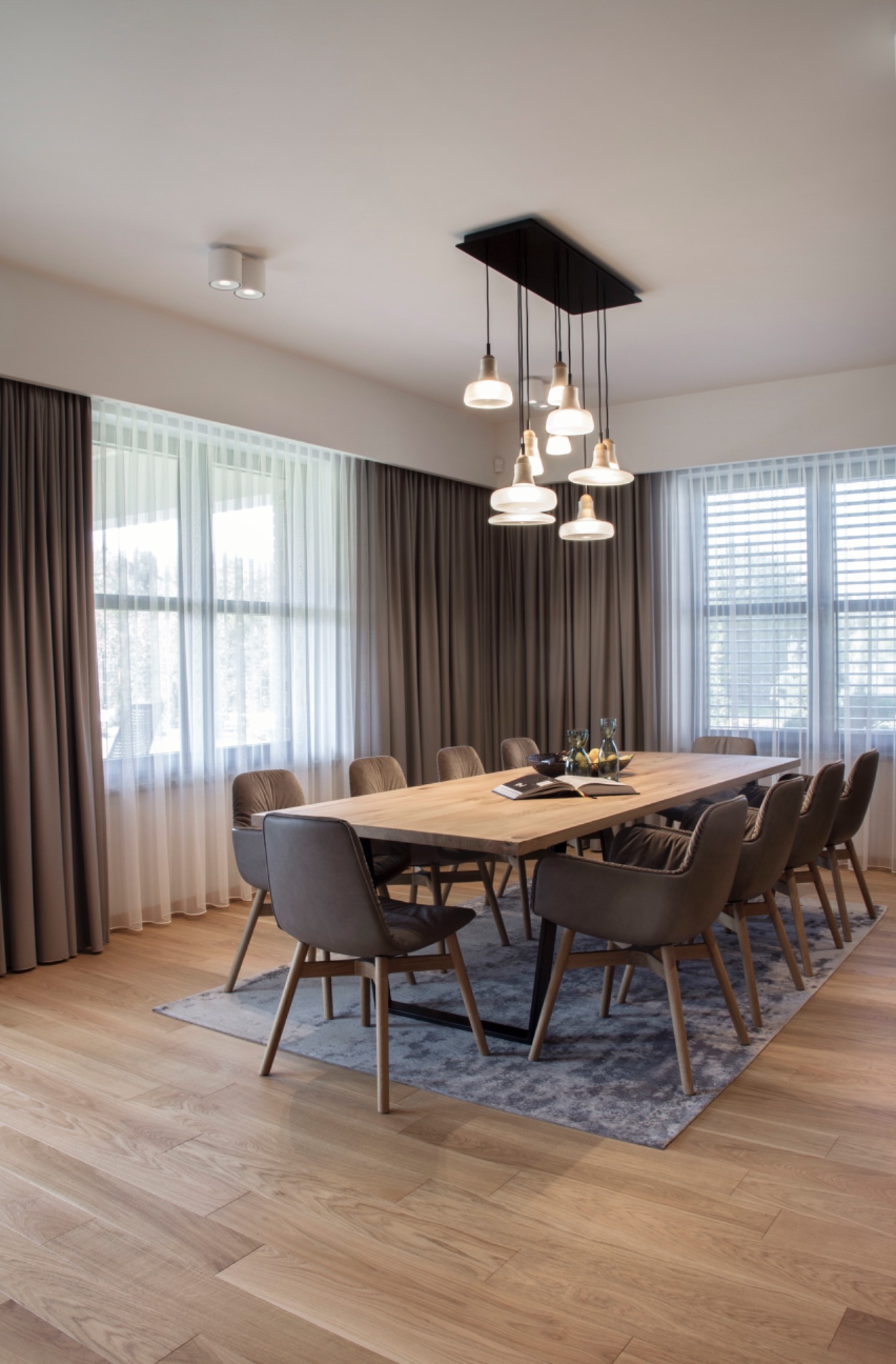 Jadalnia z długim drewnianym stołem, krzesłami obitymi skórą, szarym dywanem i zasłonami w odcieniach szarości, oświetlona wiszącą lampą z kilkoma kloszami.