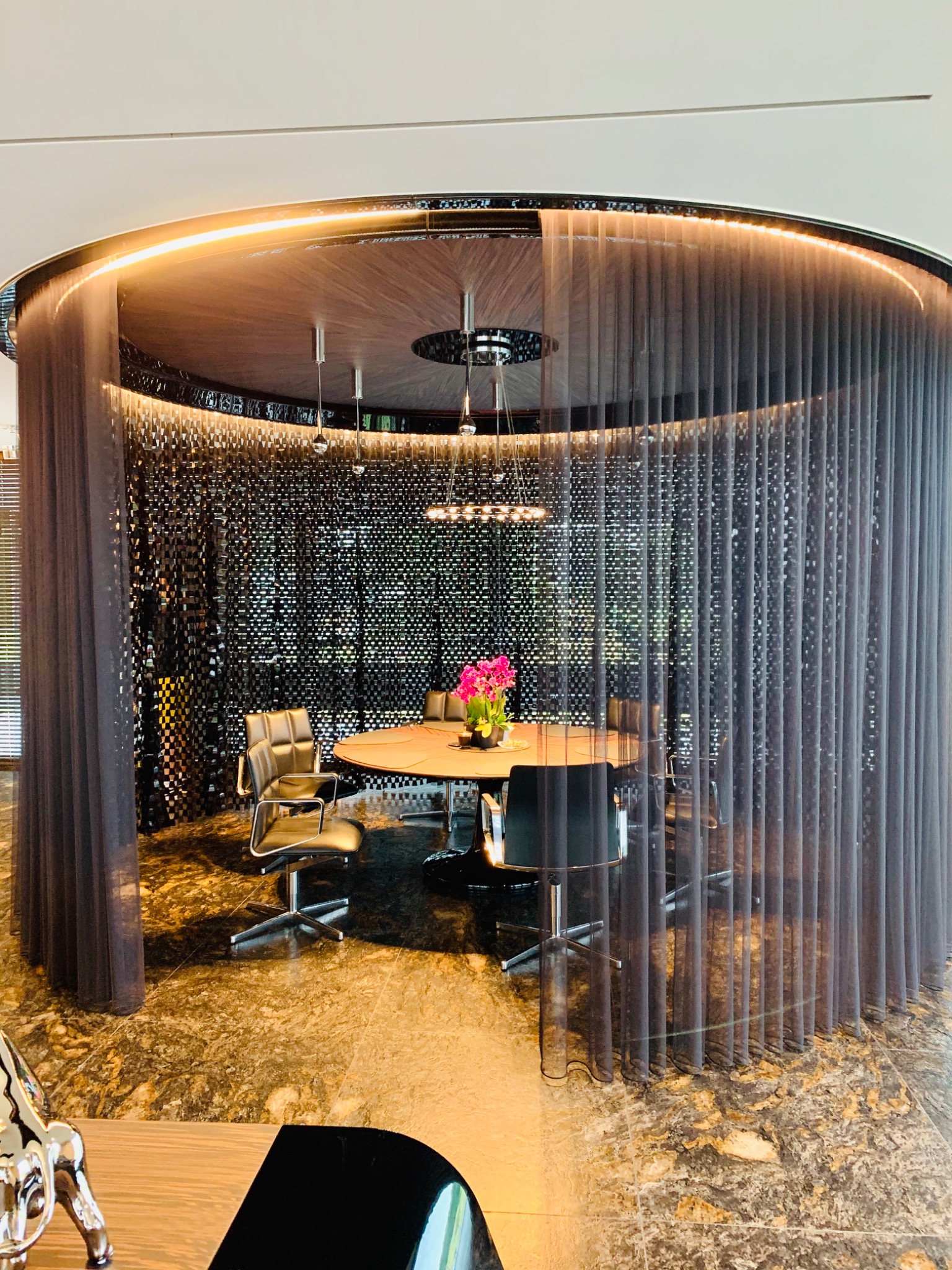 Owalny stół konferencyjny otoczony szarymi, półprzezroczystymi panelami materiału, podświetlonymi od góry, z widocznymi krzesłami biurowymi i dekoracyjnym bukietem kwiatów na stole.