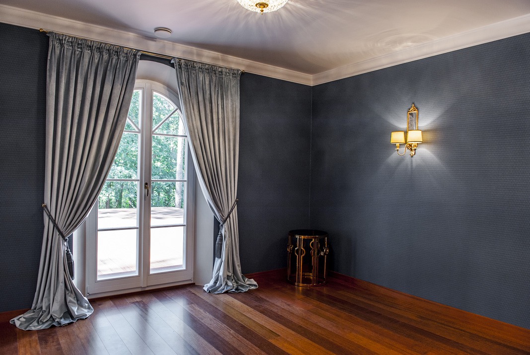 Eleganckie, drapowane zasłony w odcieniach szarości, upięte sznurami z frędzlami, zdobią półokrągłe okno w pokoju z drewnianą podłogą i ciemnoszarą ścianą z dekoracyjnym oświetleniem.