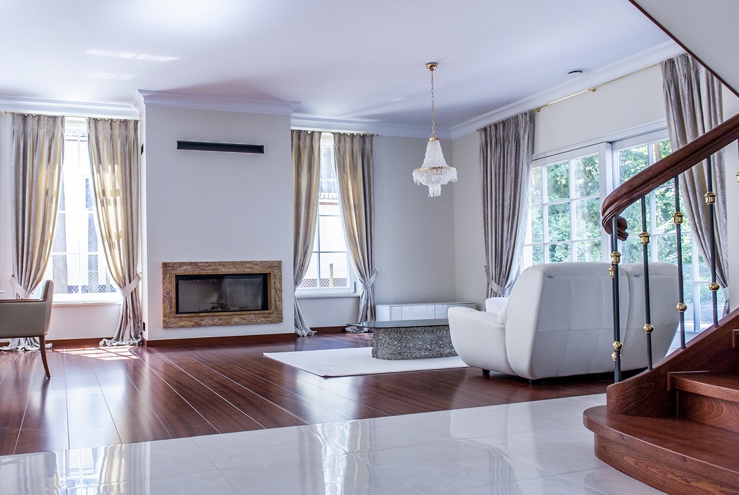 Elegancki salon z dużymi oknami, na których wiszą długie, srebrzyste zasłony, drewniana podłoga, biała sofa i kryształowy żyrandol.