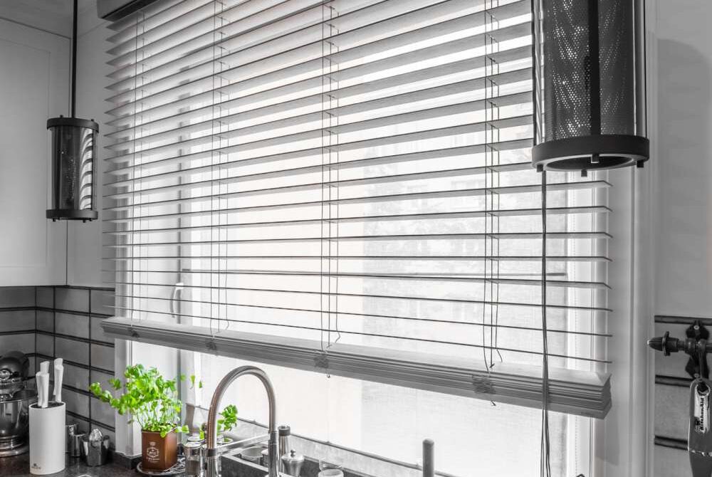 Szare żaluzje poziome zamontowane w oknie kuchennym nad zlewem, z widoczną rośliną w doniczce i lampą wiszącą w tle.