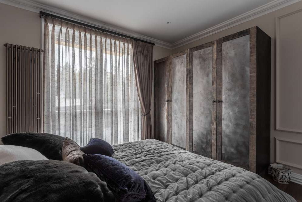 Sypialnia z szarymi, transparentnymi zasłonami woalowymi, dopasowanymi do koloru ścian i tapicerowanego łóżka, oraz wysoką, drewnianą szafą o surowym wykończeniu.