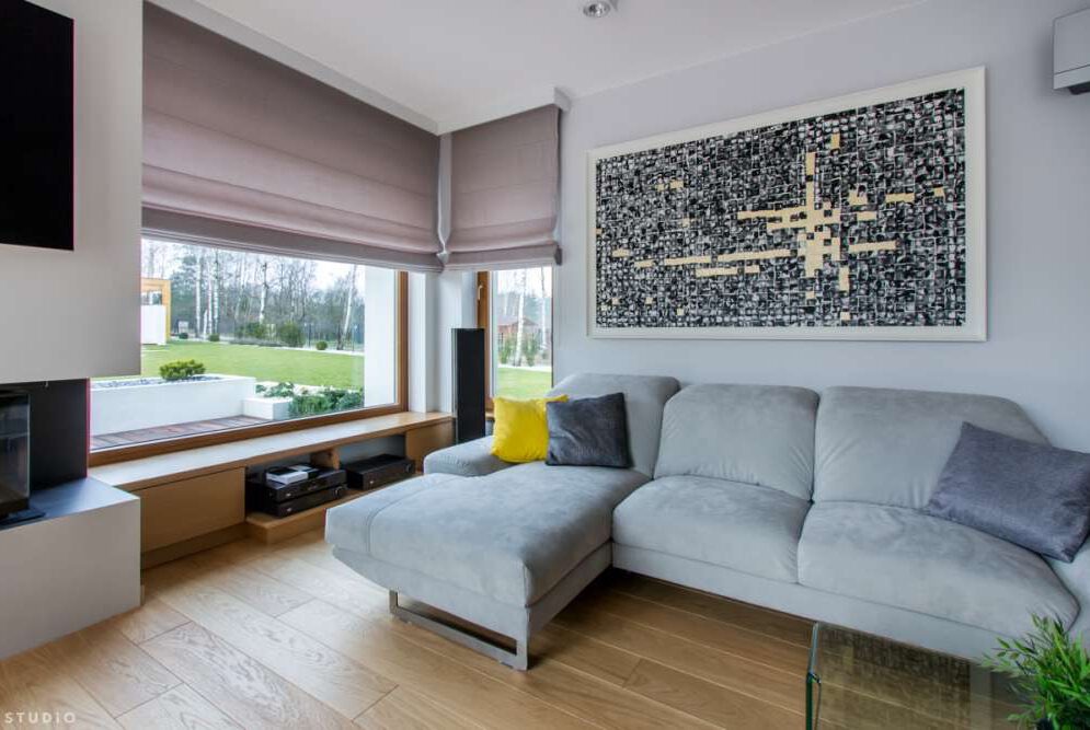 Jasnoszare rolety rzymskie zamontowane w dużym oknie salonu, widok na zielony ogród, nowoczesny wystrój wnętrza z drewnianą podłogą i szarą sofą.