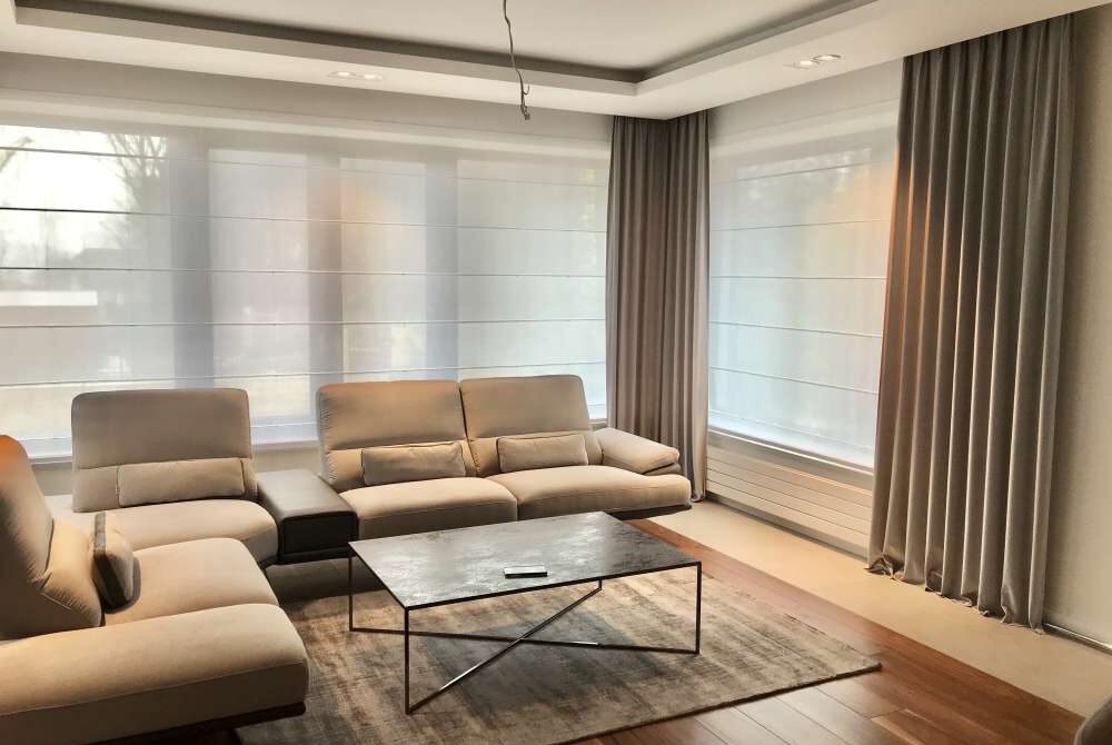 Elegancki salon z beżowymi meblami wypoczynkowymi, dywanem w odcieniach szarości, roletami rzymskimi i długimi zasłonami po bokach okien.