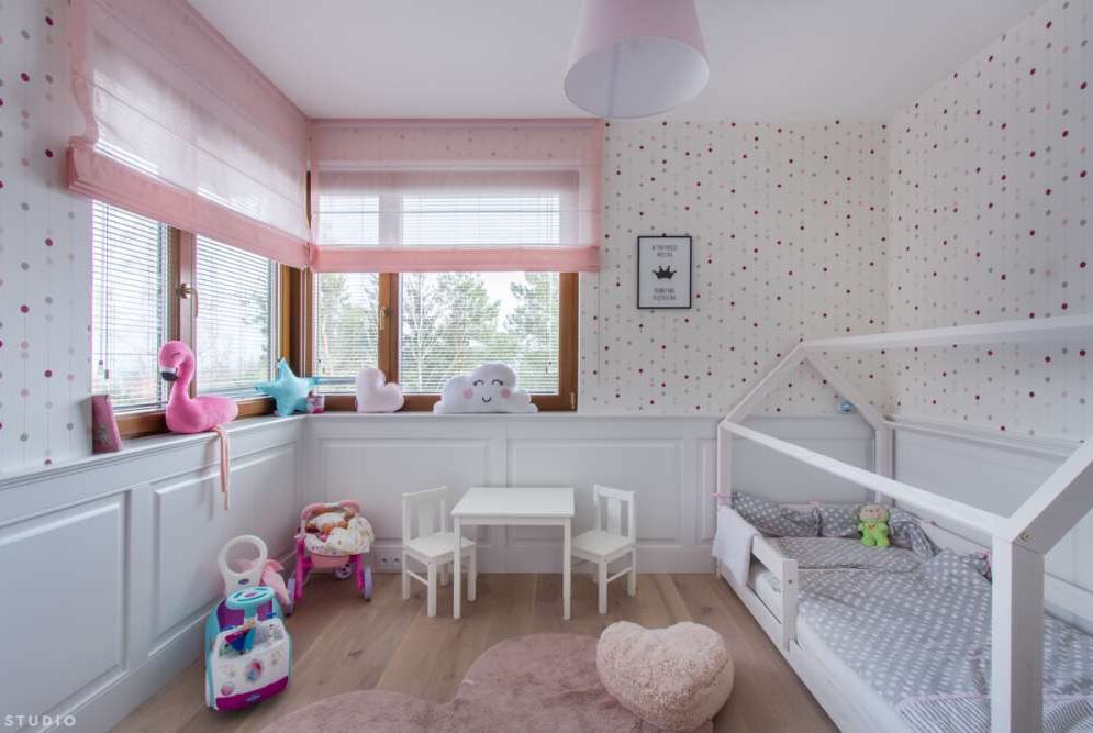 Pokój dziecięcy z różowymi roletami rzymskimi, białymi meblami i zabawkami na tle ściany w kropki.