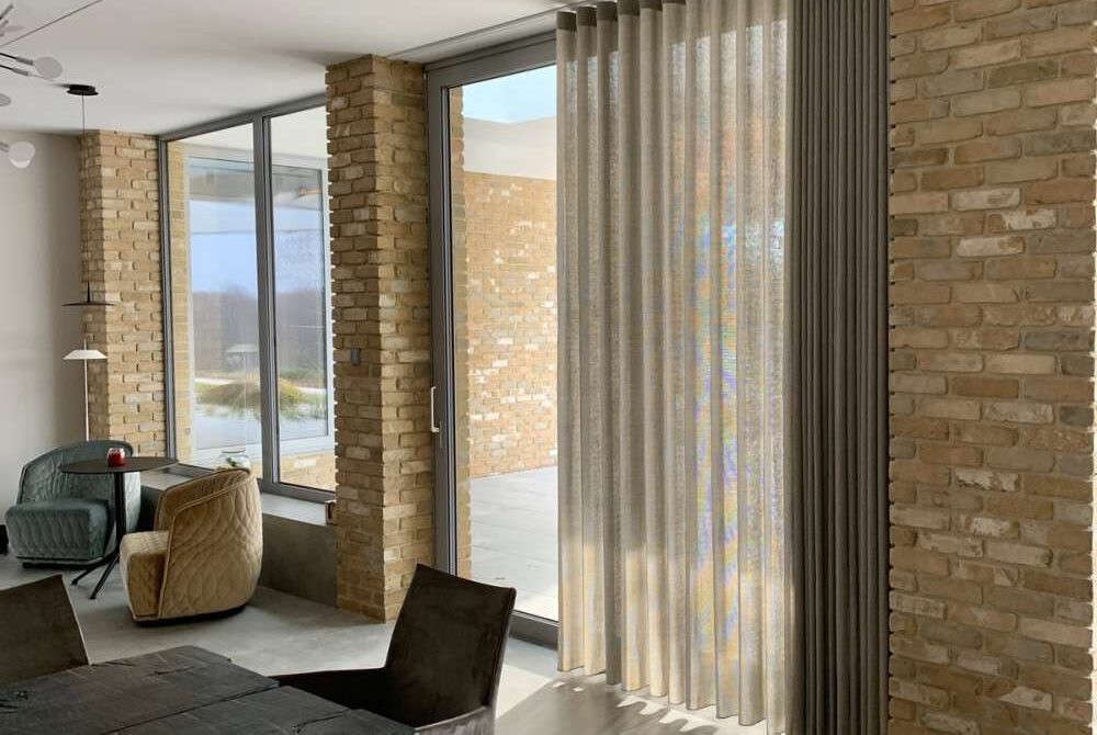 Wysokie okno z jasną, pionowo układającą się zasłoną, przylegającą do szarej, wąskiej rolety, na tle ceglanej ściany w nowoczesnym salonie.