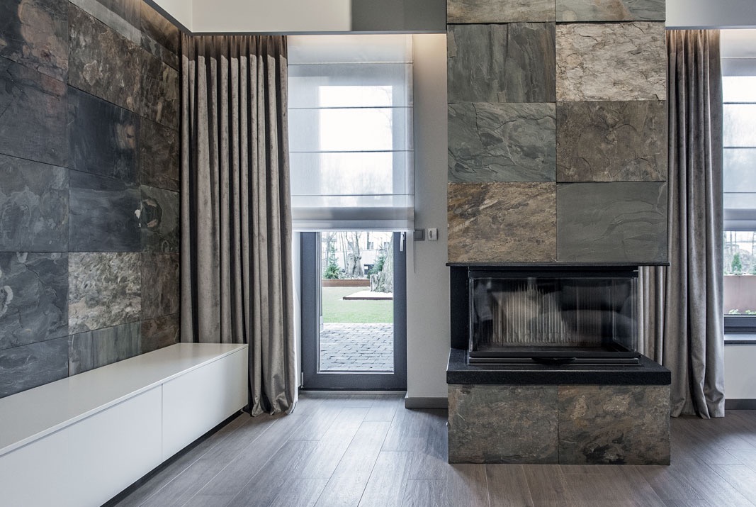 Nowoczesny salon z kamienną ścianą, kominkiem obłożonym płytami i szarymi, długimi zasłonami, uzupełniony roletą rzymską w oknie balkonowym.