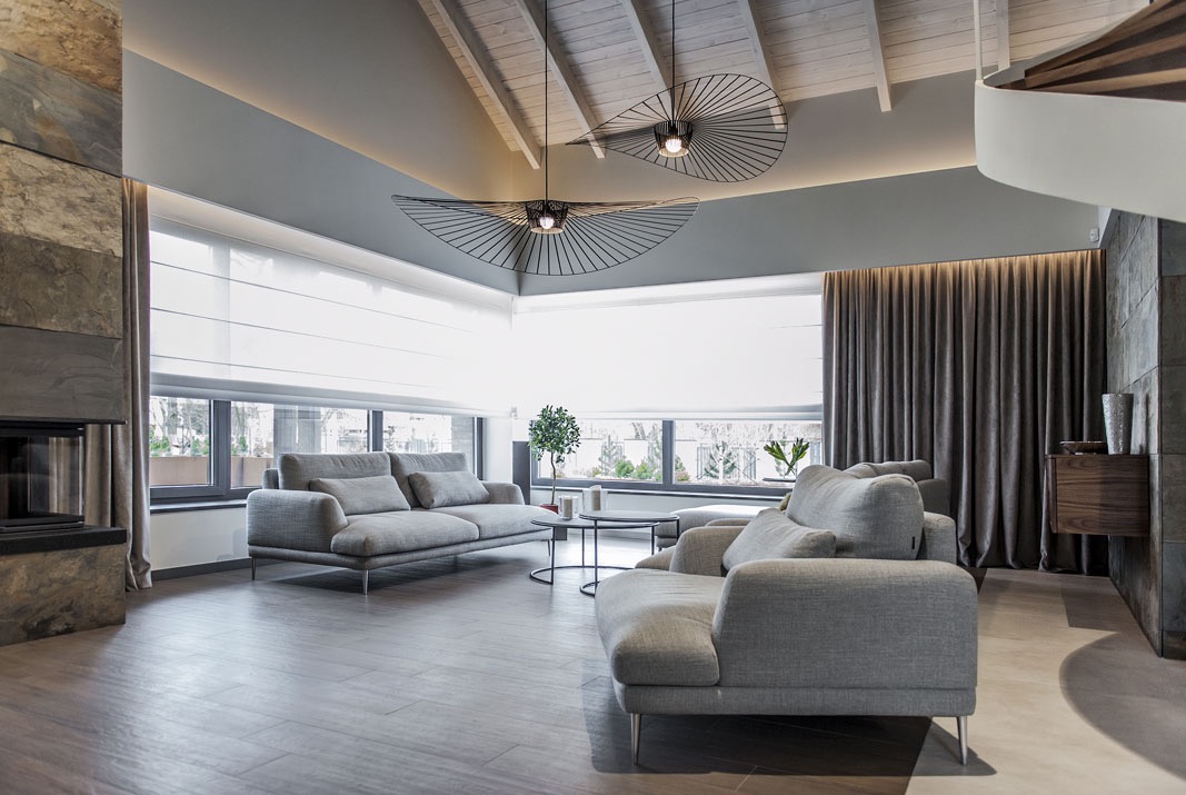 Jasny salon z szarymi sofami, drewnianą podłogą, oknami z roletami w kasecie i oryginalnymi lampami sufitowymi w kształcie wachlarzy.