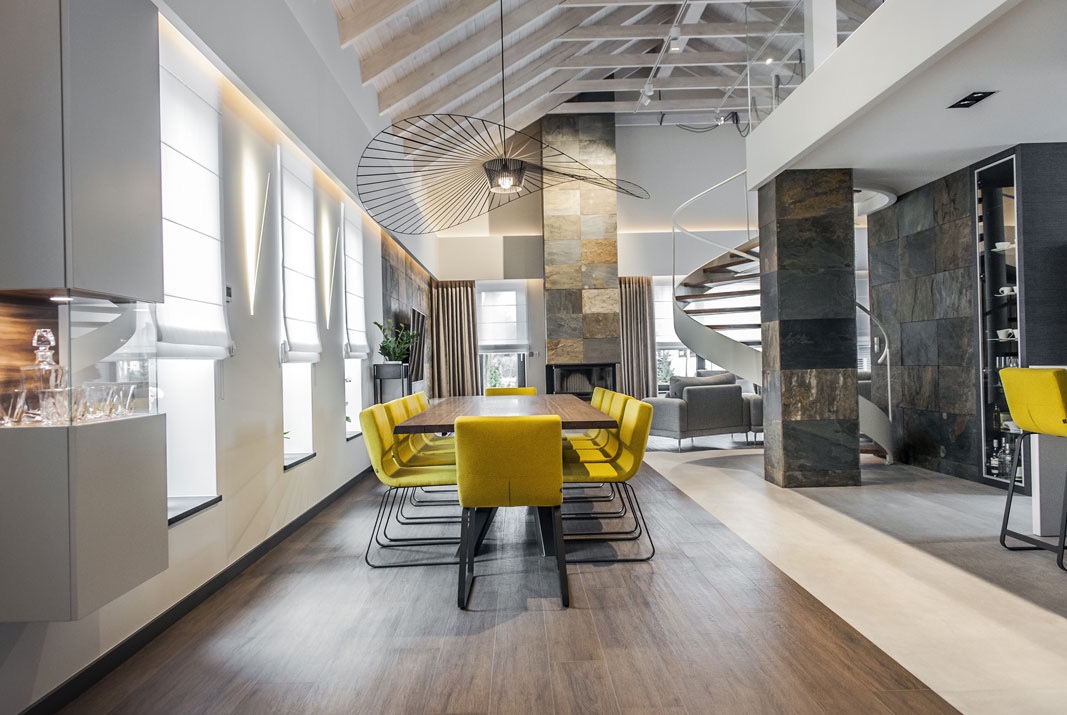 Eleganckie wnętrze jadalni z białymi roletami rzymskimi w oknach, żółtymi krzesłami przy drewnianym stole, spiralnymi schodami i dekoracyjnym żyrandolem.