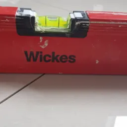 Czerwona poziomica marki Wickes leży na jasnych, świeżo położonych płytkach podłogowych, widoczne ślady zaprawy.