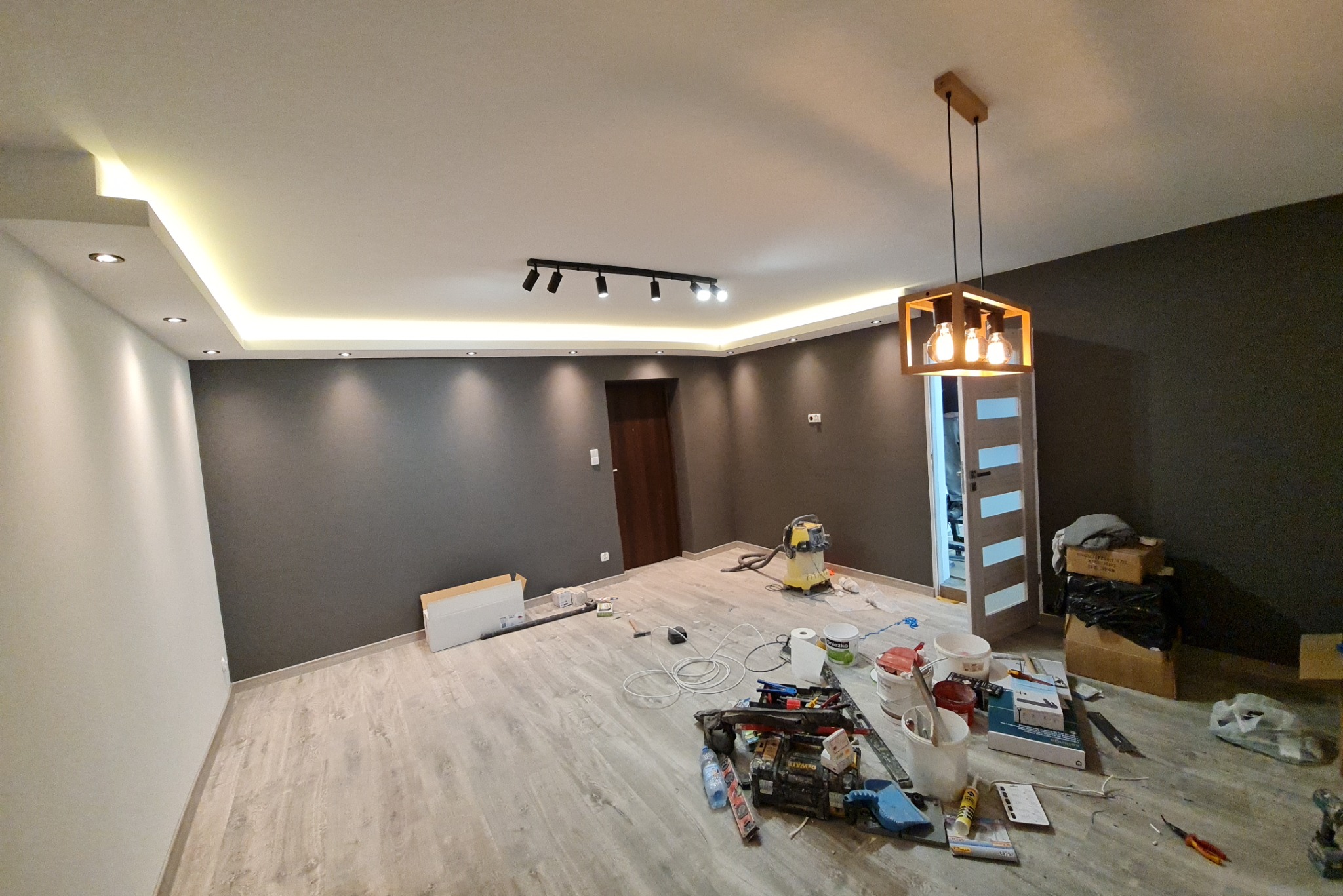 Pokój w trakcie remontu: szare ściany, jasna podłoga, oświetlenie sufitowe LED, wisząca lampa z drewnianą ramą, narzędzia i materiały malarskie rozłożone na podłodze, odkurzacz przemysłowy w tle.