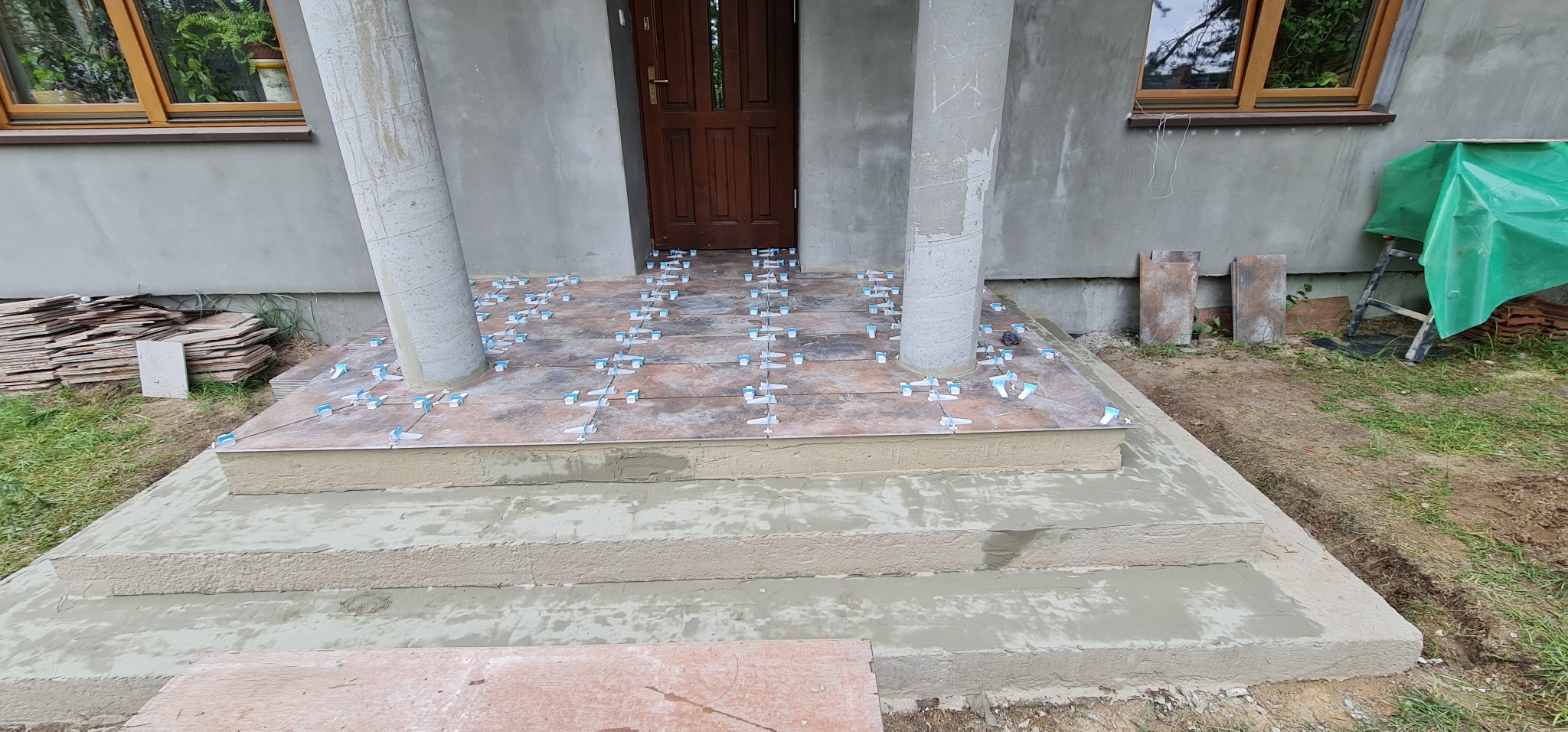 Montaż płytek imitujących drewno na schodach wejściowych do domu, z użyciem poziomujących krzyżyków dystansowych i zaprawy, widoczne kolumny i okna budynku.