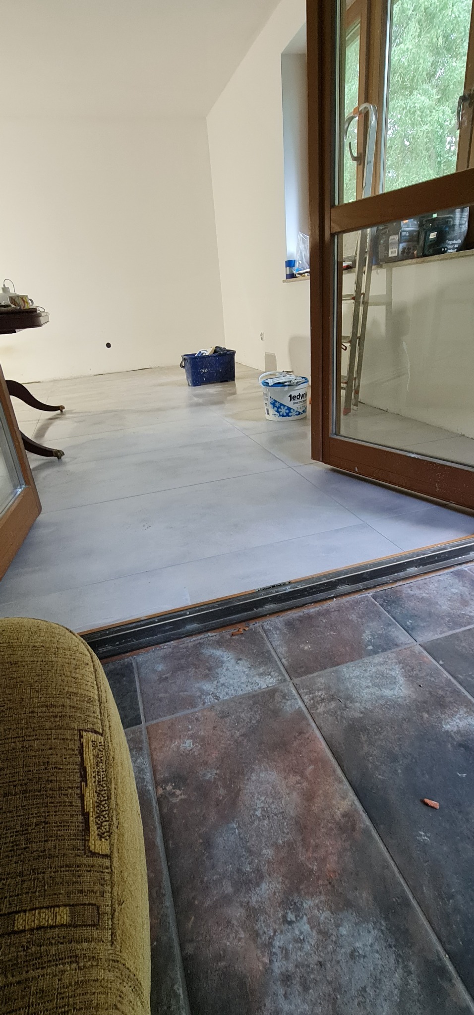 Wnętrze w trakcie malowania ścian na biało i układania jasnych płytek podłogowych, widoczne wiadro z farbą i kuweta malarska, z fragmentem brązowych płytek na pierwszym planie.