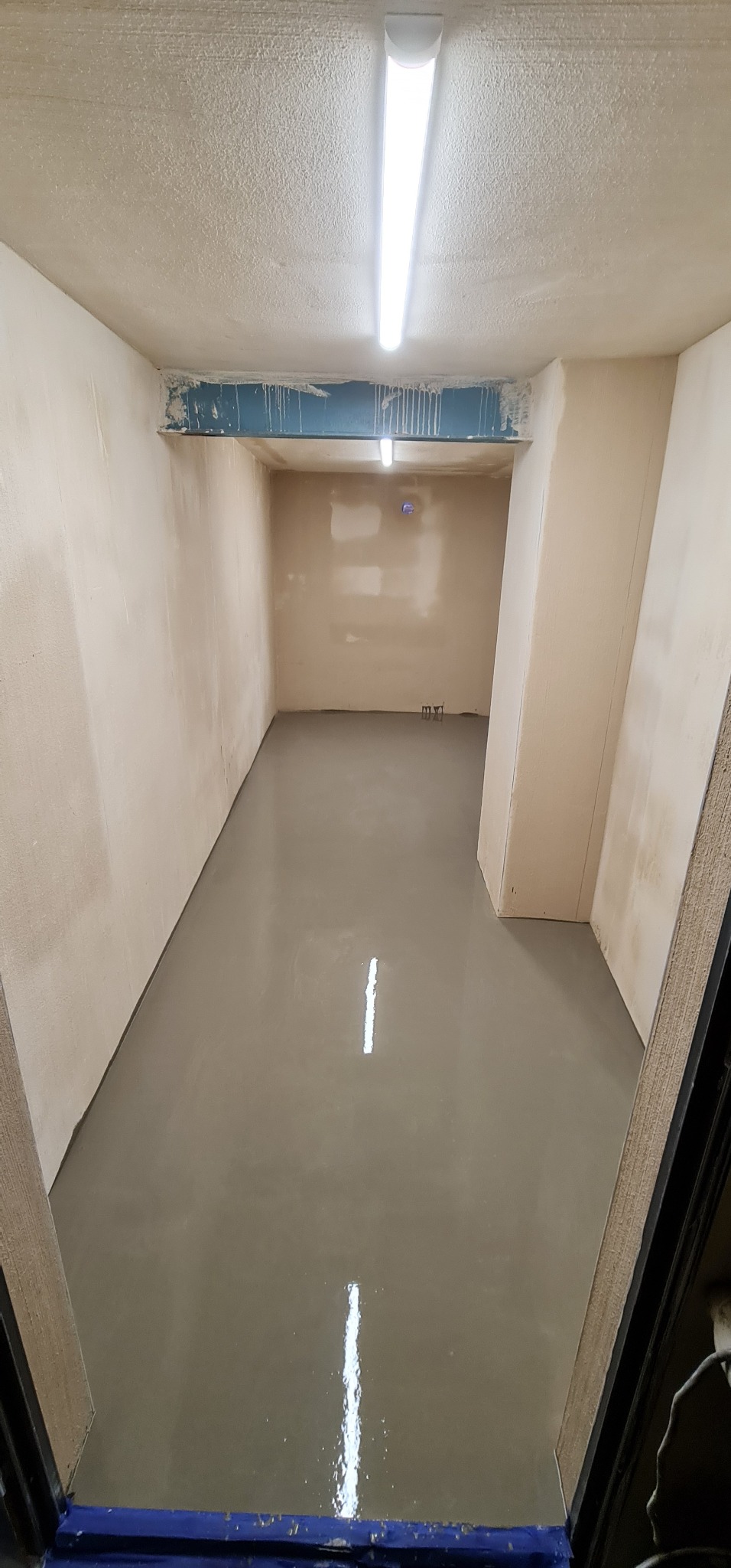 Wylewka samopoziomująca na podłodze w pomieszczeniu w trakcie prac wykończeniowych, widoczne ślady po malowaniu na ścianach i suficie, oświetlenie jarzeniowe.
