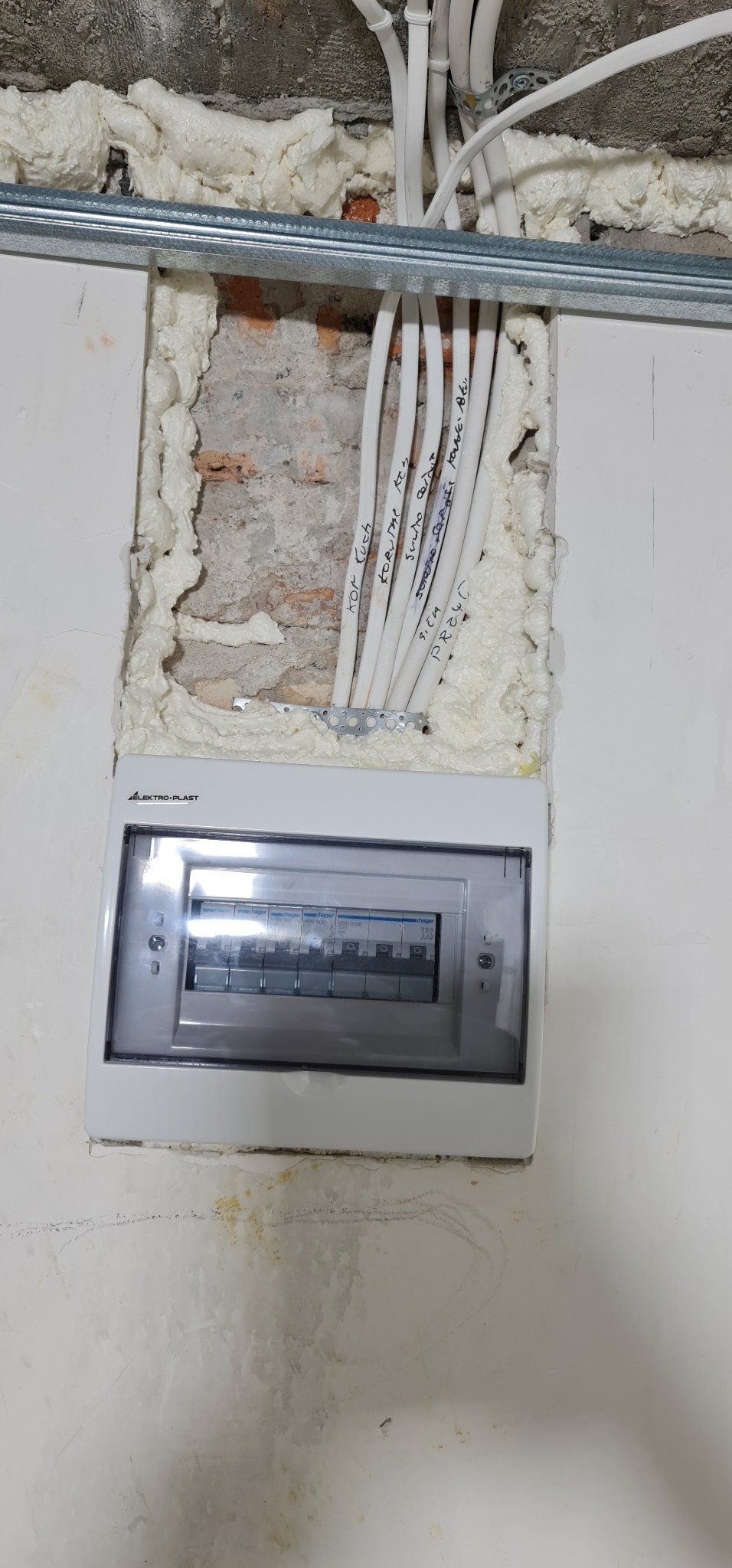 Rozdzielnica elektryczna Elektro-Plast zamontowana w ścianie z widocznymi przewodami oznaczonymi napisami, zabezpieczonymi pianką montażową i osadzonymi w otworze w ceglanej ścianie.