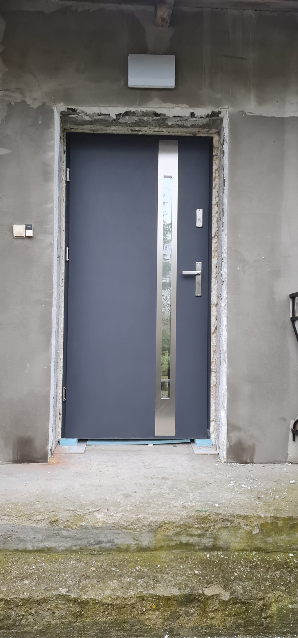 Montaż nowoczesnych, grafitowych drzwi zewnętrznych z pionowym przeszkleniem na tle surowego muru z widocznymi śladami zaprawy i pianki montażowej.