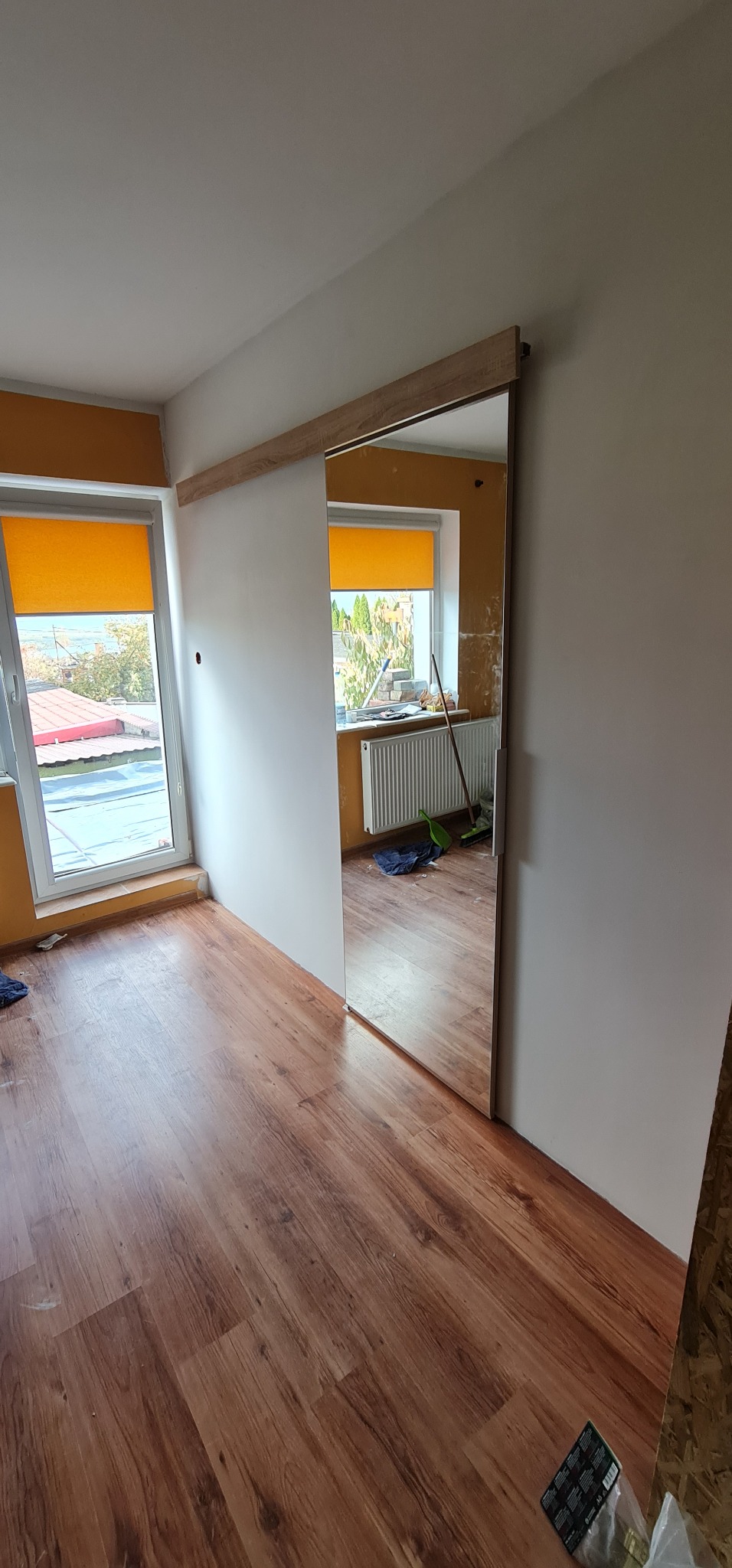 Montaż przesuwnych drzwi z drewnianym prowadzeniem w odnawianym pokoju z panelami podłogowymi, widoczne okna z żółtymi roletami i grzejnik.