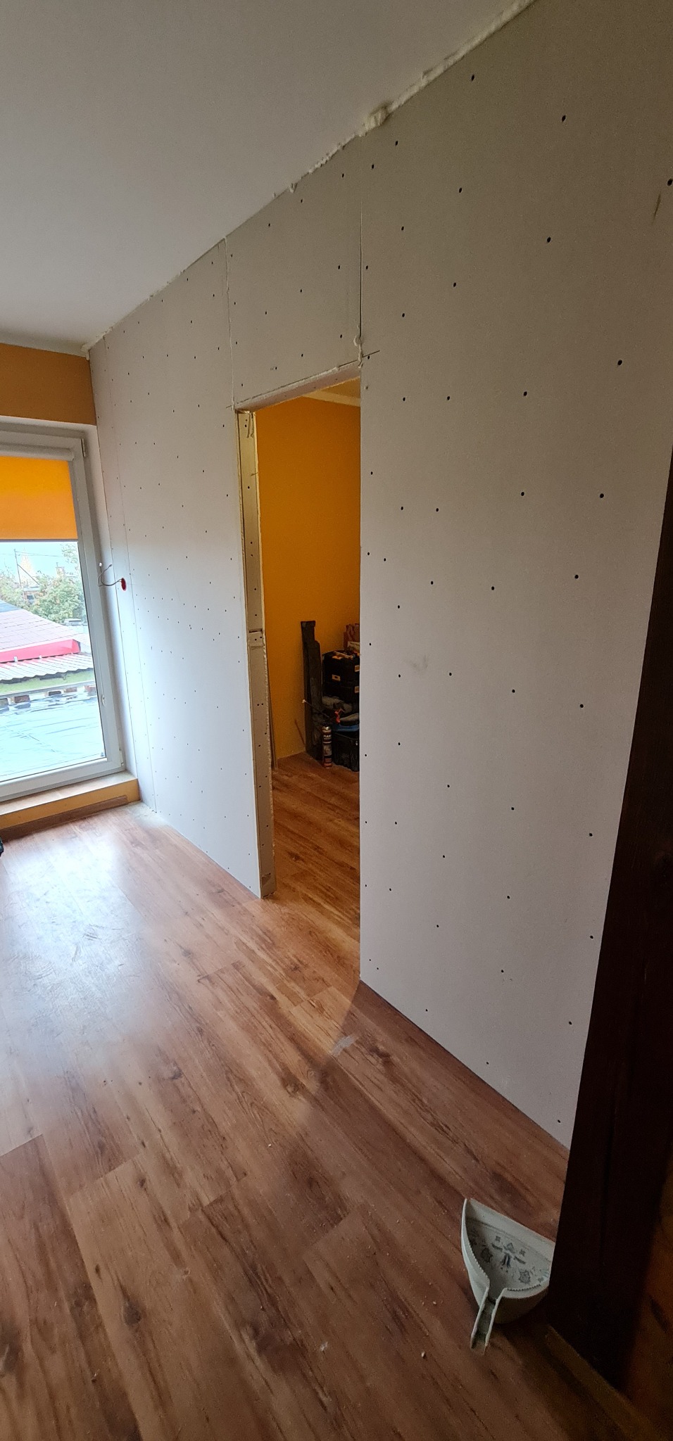 Montaż płyt karton gips na ścianie w pokoju z oknem i drewnianą podłogą; widoczny otwór drzwiowy i narzędzie do szpachlowania na podłodze.