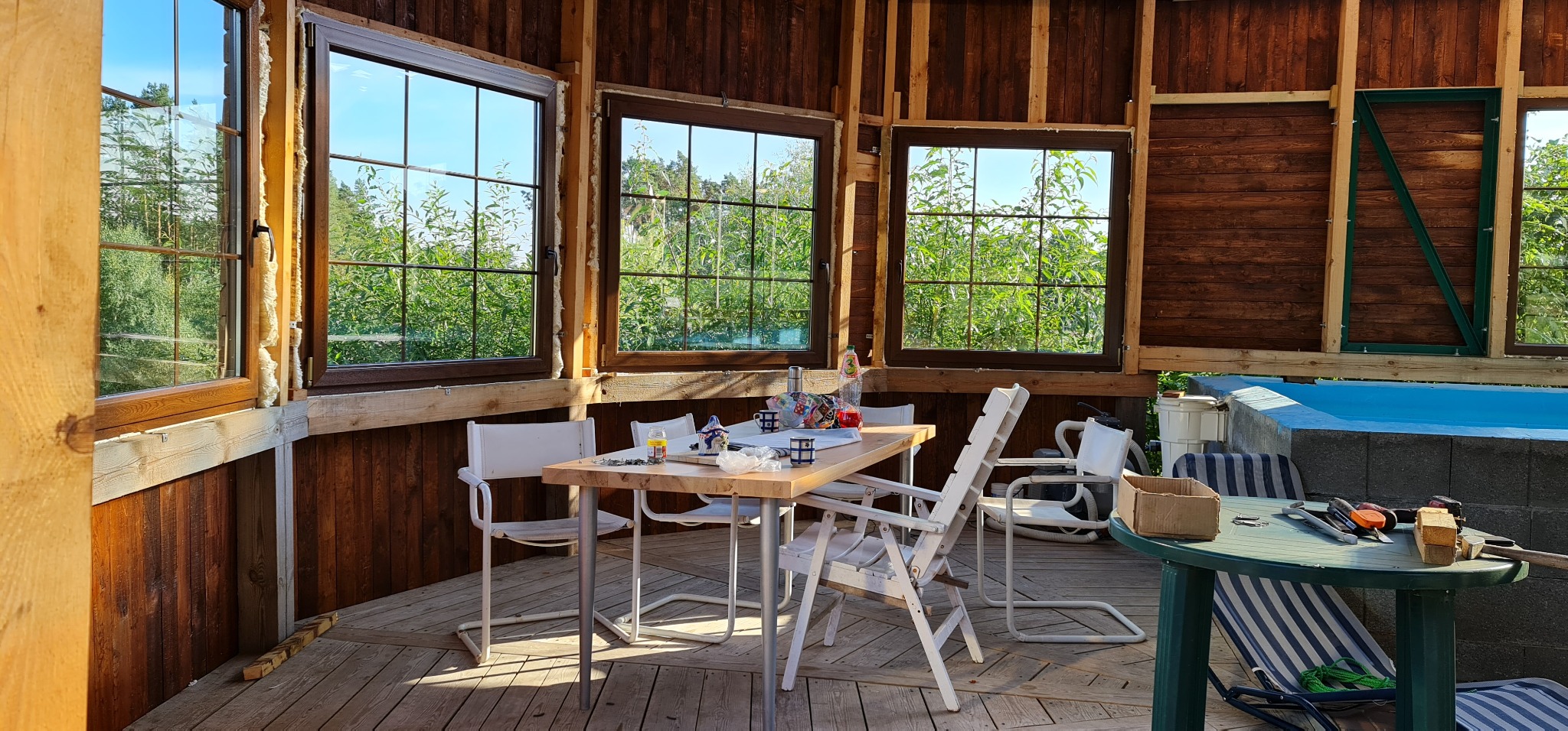 Wnętrze drewnianej altany z częściowo zamontowanymi oknami z widokiem na zieleń, stół z krzesłami w centralnej części, w tle widoczny basen w trakcie budowy.