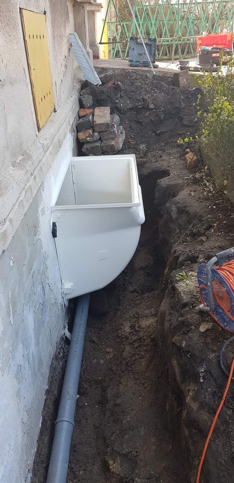 Montaż białego okna piwnicznego z rurą odpływową w wykopie przy ścianie budynku z żółtymi drzwiami i stertą cegieł.