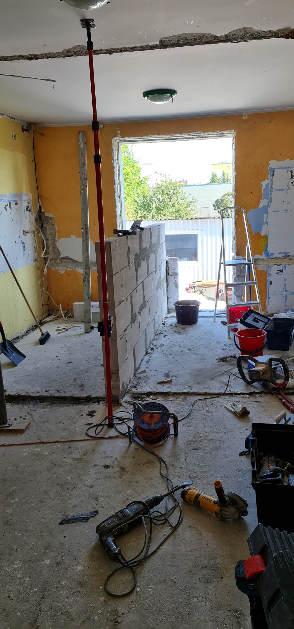 Wnętrze w trakcie budowy ściany działowej z bloczków, widoczne narzędzia budowlane, poziomica laserowa oparta o sufit, ściany w stanie surowym z resztkami farby.