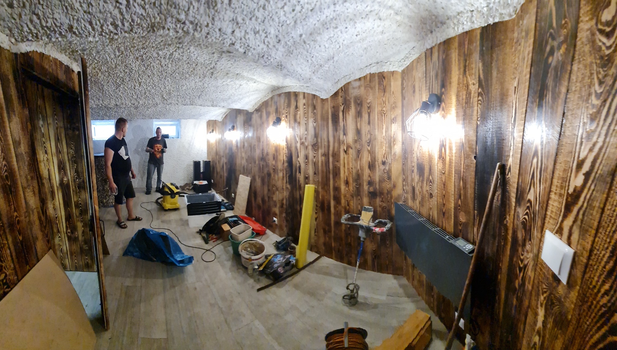 Piwnica w trakcie prac wykończeniowych: drewniana boazeria na ścianach, sufit pokryty białą pianką, widoczne narzędzia i materiały budowlane, dwóch mężczyzn w tle.