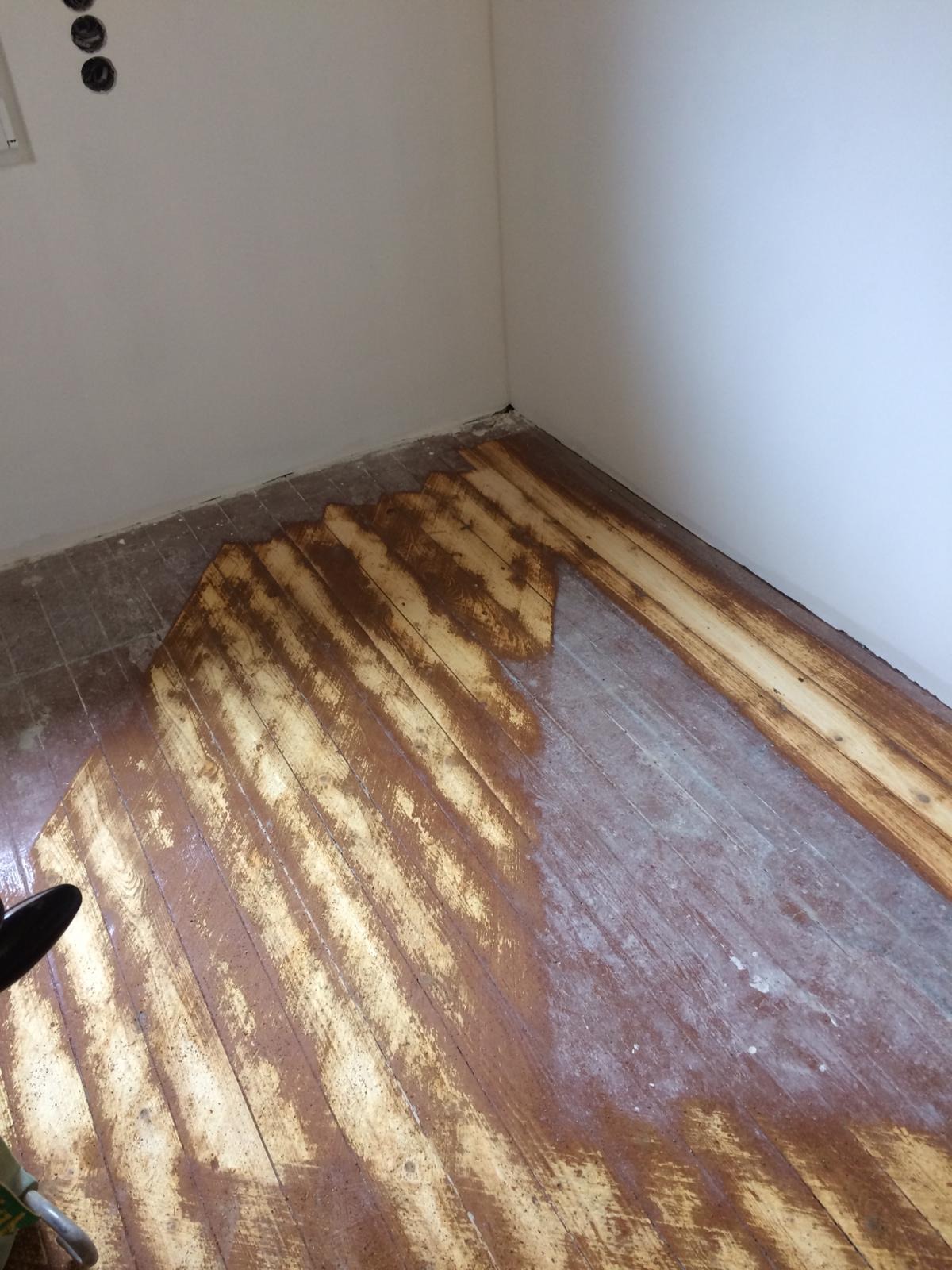 Fragment pokoju w trakcie renowacji podłogi drewnianej; widoczne deski podłogowe częściowo pokryte szlifowaną warstwą, z widocznymi śladami po cykliniarce.