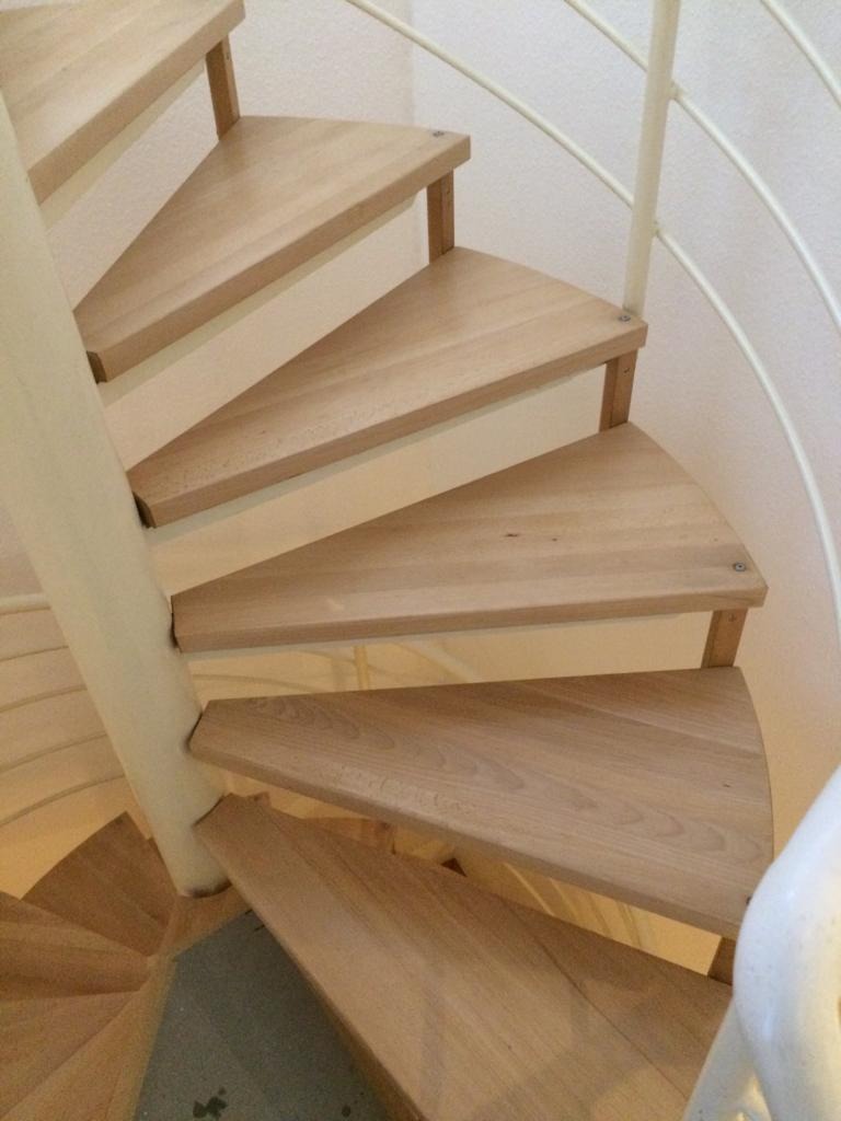 Schody spiralne z jasnego drewna, widok z góry na kilka stopni, biała metalowa balustrada.