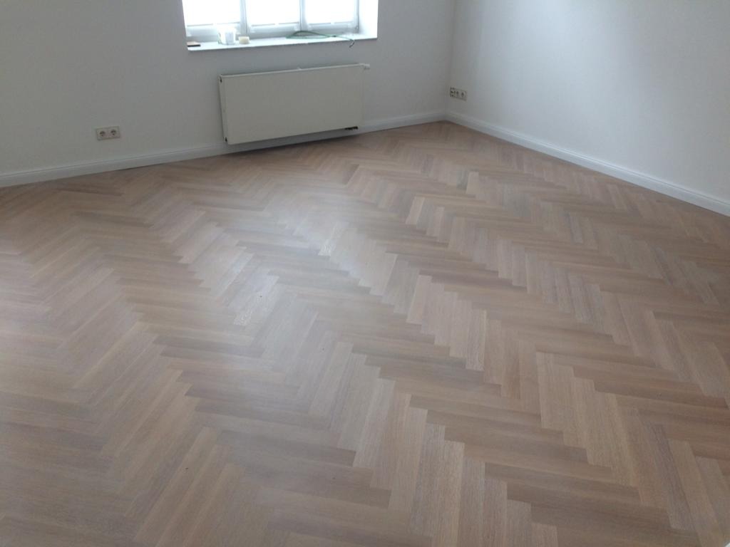 Jasna, nowo ułożona podłoga drewniana w jodełkę klasyczną, widok z góry na puste, białe wnętrze z grzejnikiem przy oknie.