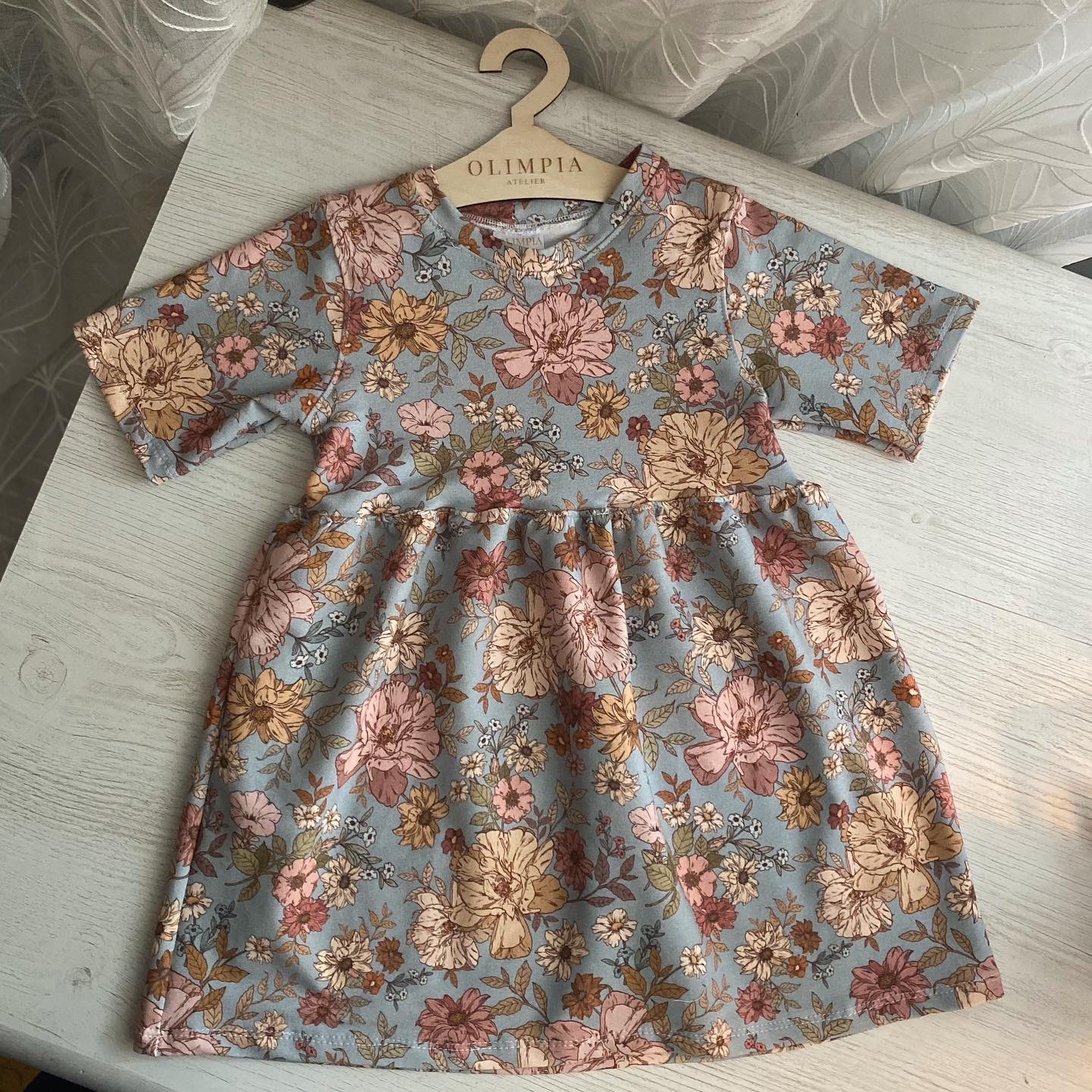 Dziecięca sukienka z krótkim rękawem w kwiatowy wzór, zawieszona na drewnianym wieszaku z napisem 'OLIMPIA ATELIER', leżąca na jasnym blacie.