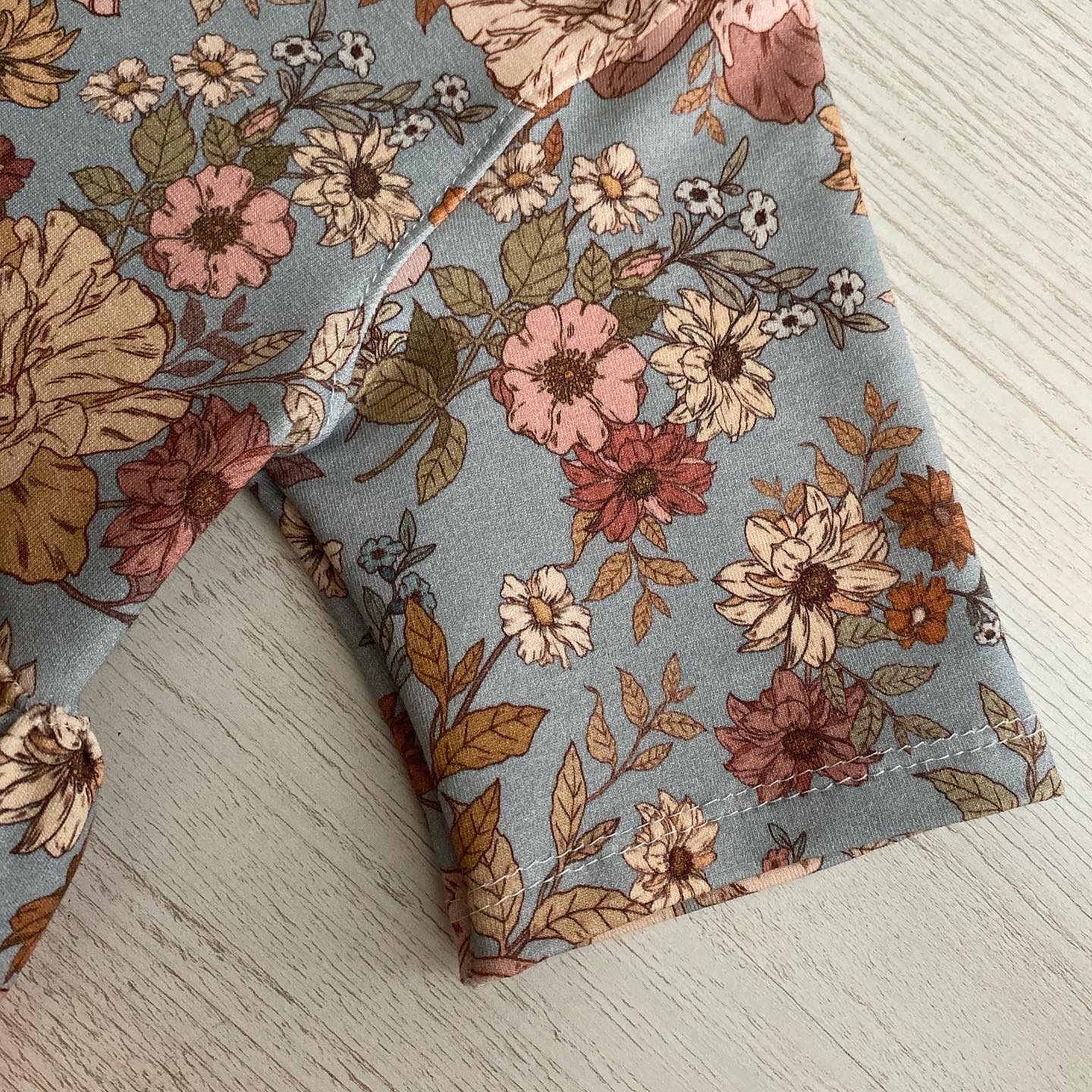 Fragment rękawa dziecięcego pajacyka z bawełny w pastelowe kwiaty na jasnym drewnianym tle, widoczne szwy wykończeniowe.