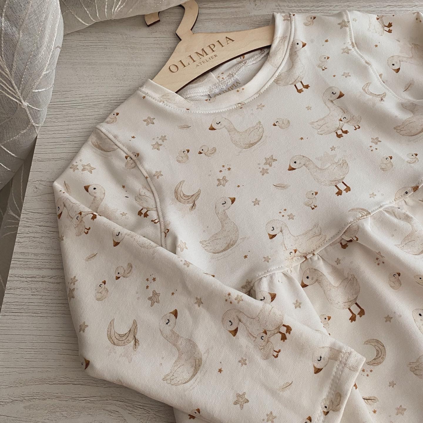 Biała sukienka dziecięca z motywem gęsi i gwiazdek, zawieszona na drewnianym wieszaku z napisem 'OLIMPIA ATELIER', leżąca na jasnym drewnianym blacie, z delikatną zasłoną w tle.