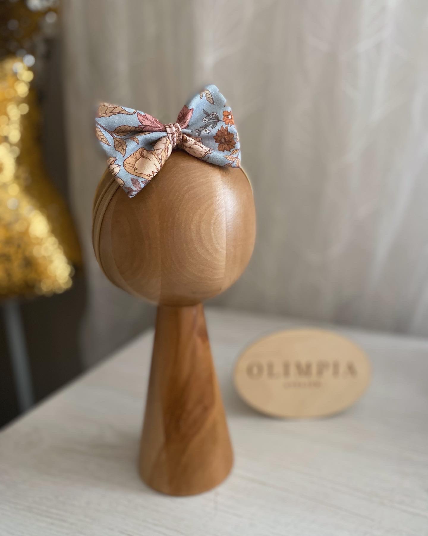 Ręcznie robiona opaska do włosów z kokardą w kwiatowy wzór, eksponowana na drewnianym stojaku w Bystrzycy, z rozmytym złotym manekinem w tle.