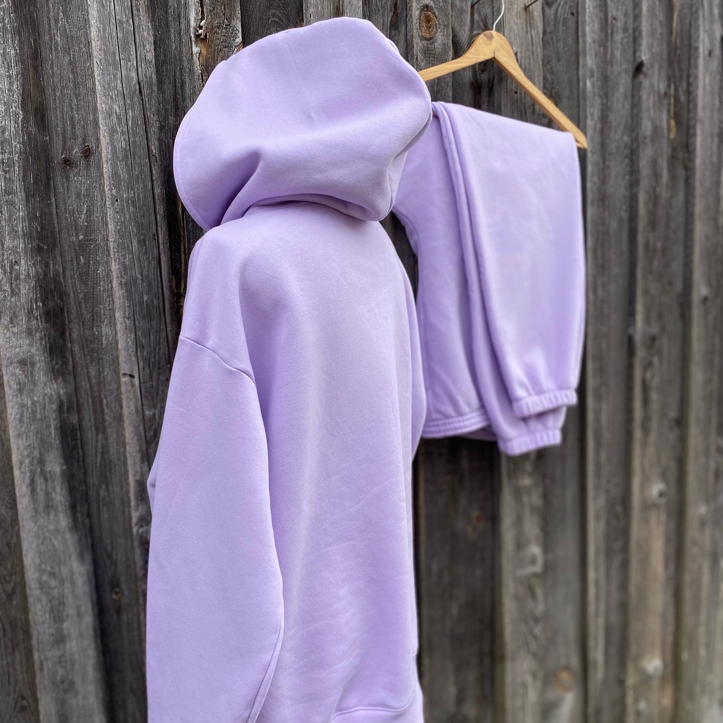 Liliowy dres damski z kapturem, bluza i spodnie dresowe, wiszące na drewnianym wieszaku na tle szarej, drewnianej ściany.