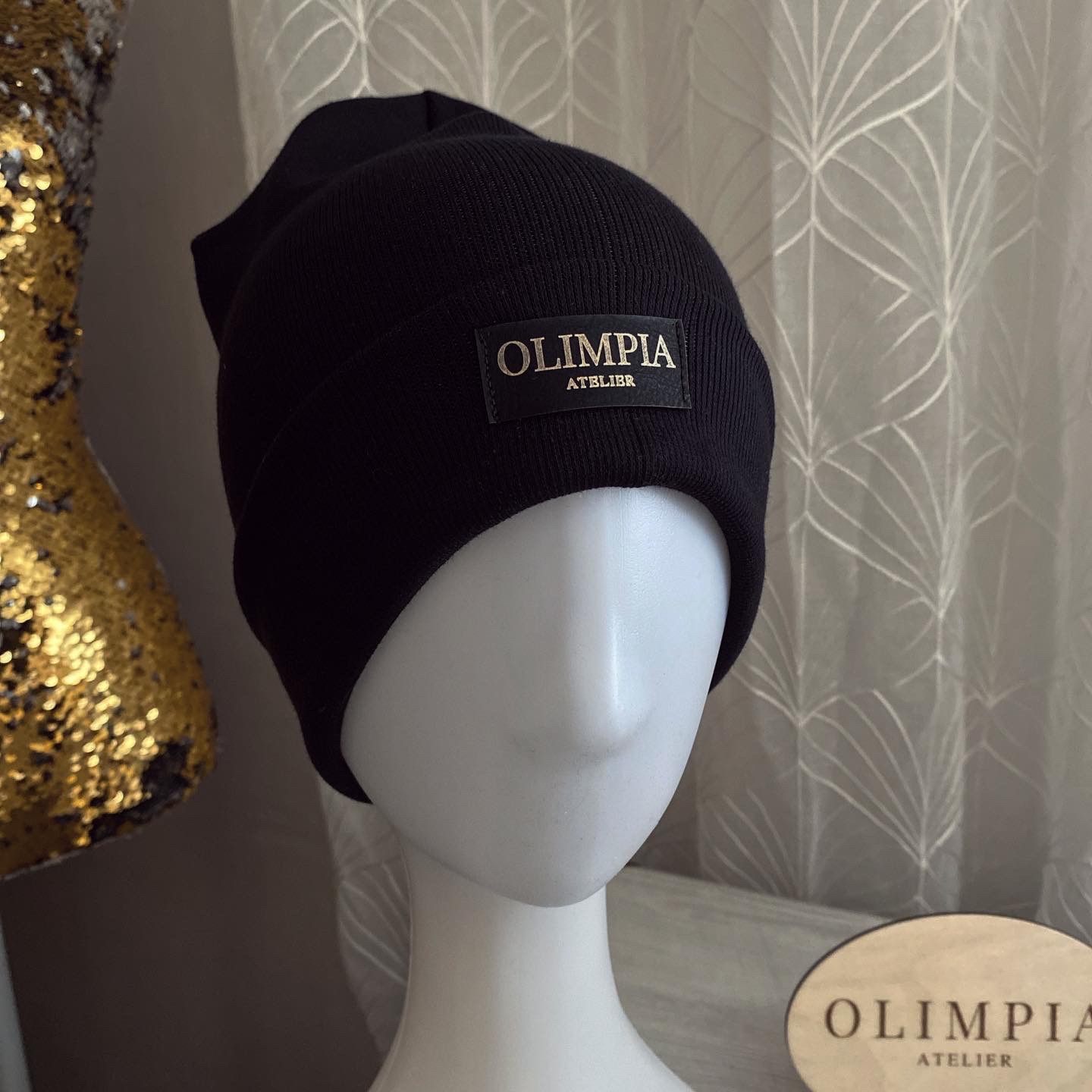 Czarna dzianinowa czapka z podwinięciem, z naszywką z napisem 'OLIMPIA ATELIER', prezentowana na białym manekinie, obok złotej, cekinowej dekoracji i drewnianego logo.