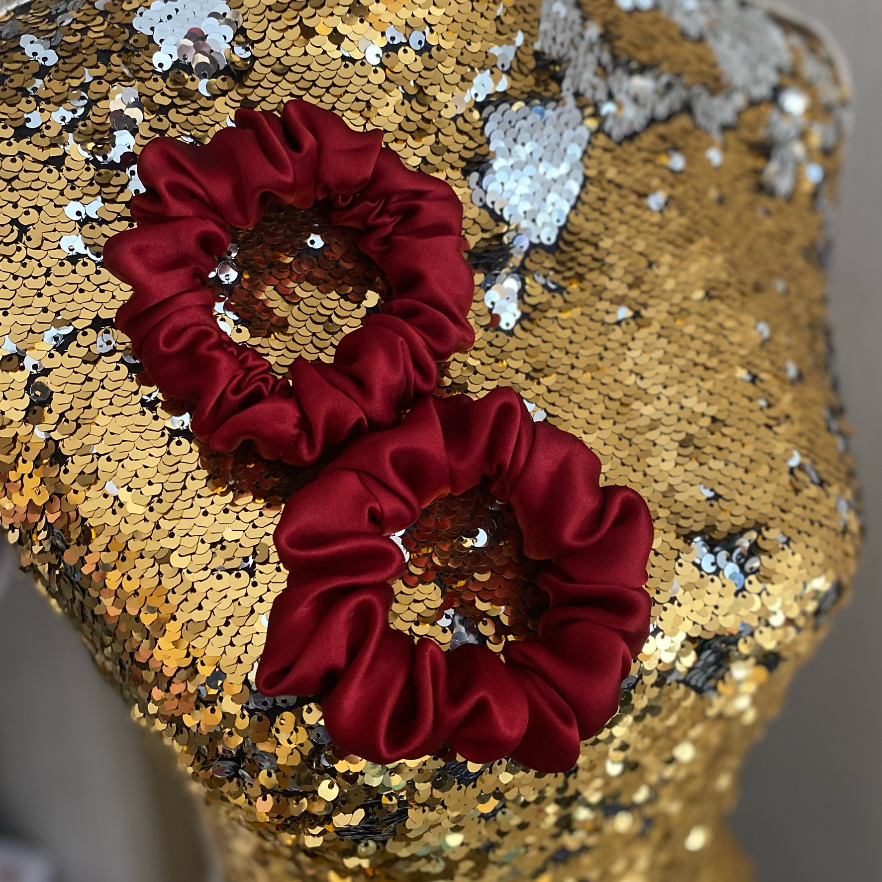 Dwie bordowe gumki do włosów z satynowego materiału na złotym, cekinowym manekinie.